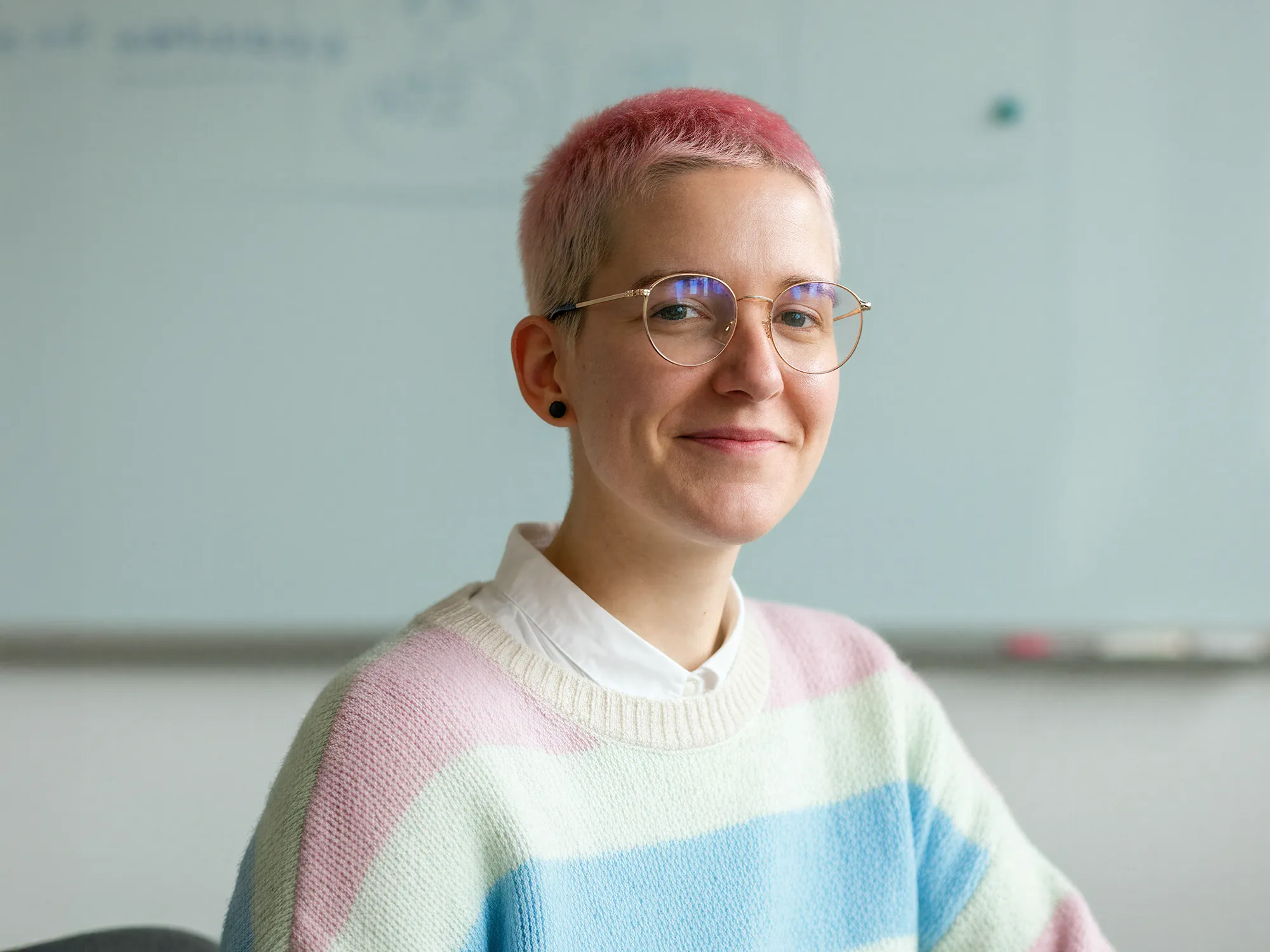 Young adult with short pink hair and glasses smiling, wearing a pastel striped sweater and white collared shirt.