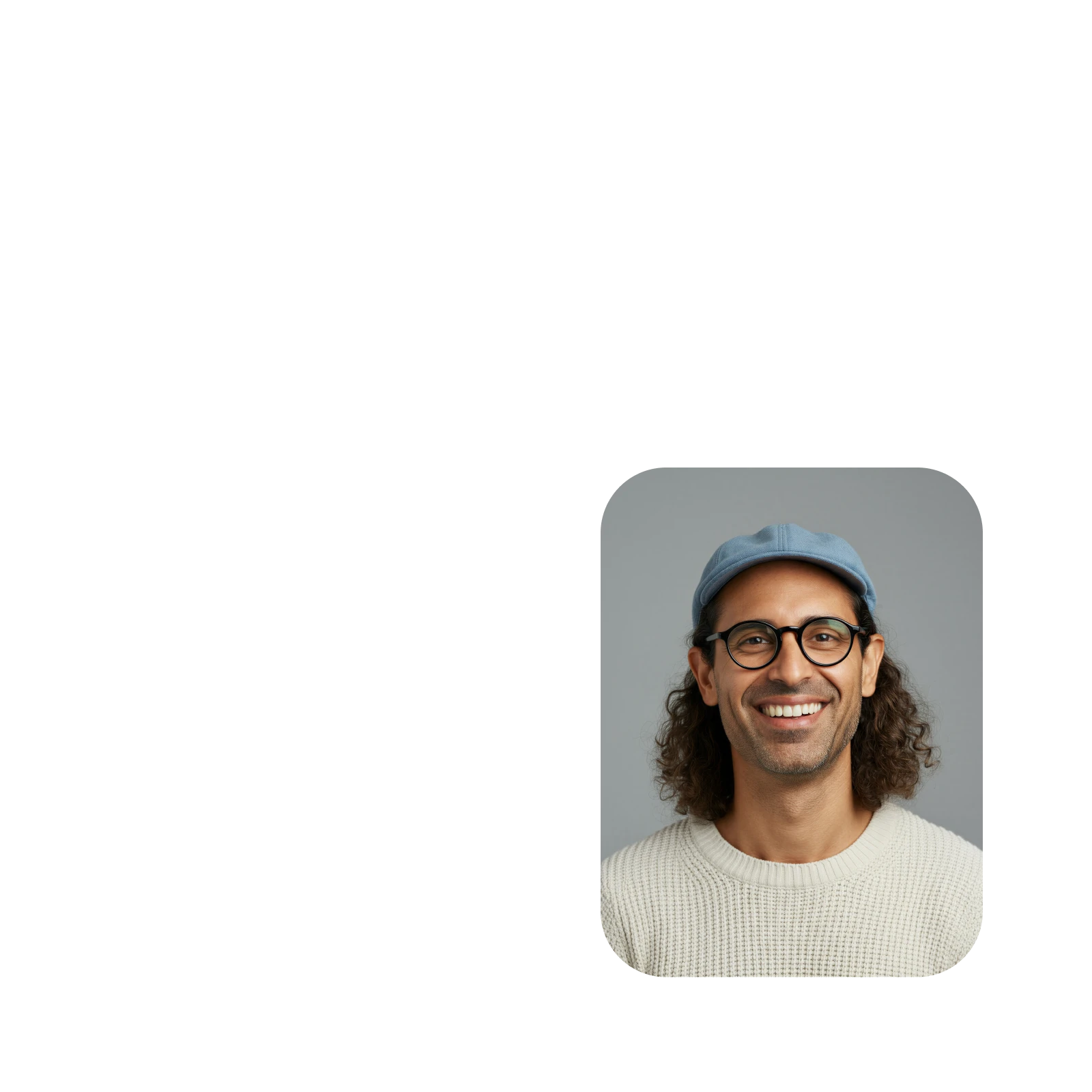 Smiling man with curly hair wearing glasses, a blue cap, and a cream knitted sweater against a gray background.