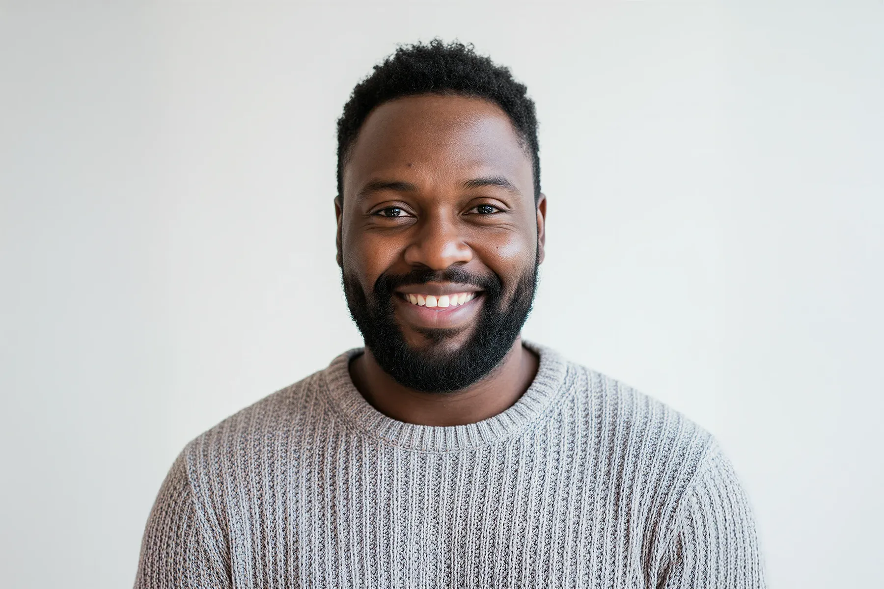 Smiling man with a beard wearing a gray knitted sweater against a plain light background.