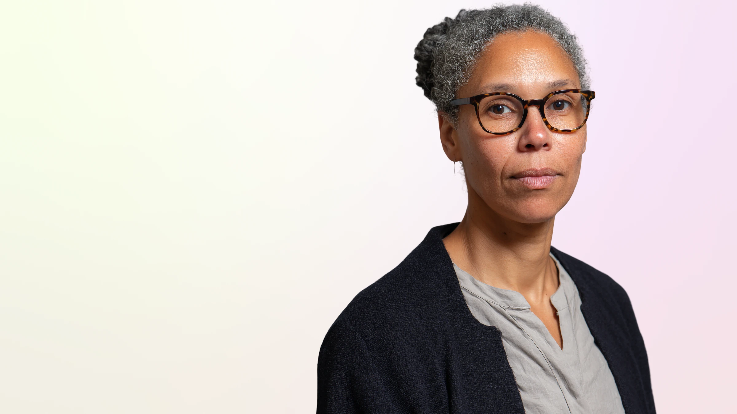 Portrait of a woman with curly gray hair in a bun, wearing tortoiseshell glasses, a gray blouse, and a black cardigan against a soft gradient background.