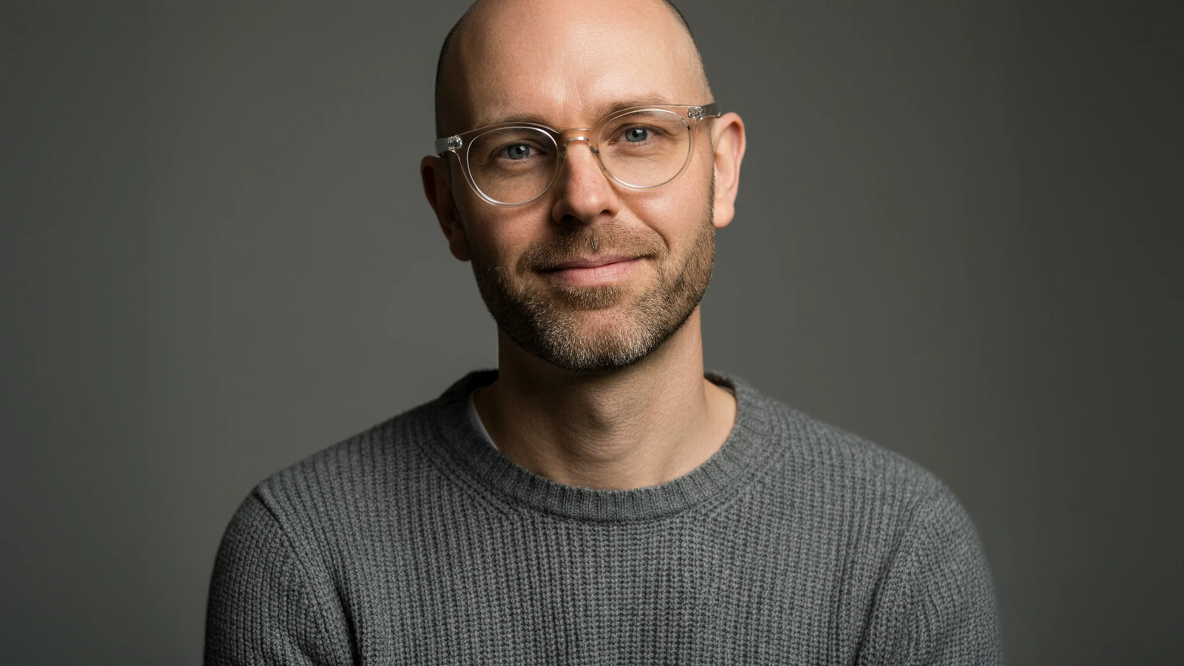 Smiling bald man with glasses wearing a gray knit sweater against a dark background.
