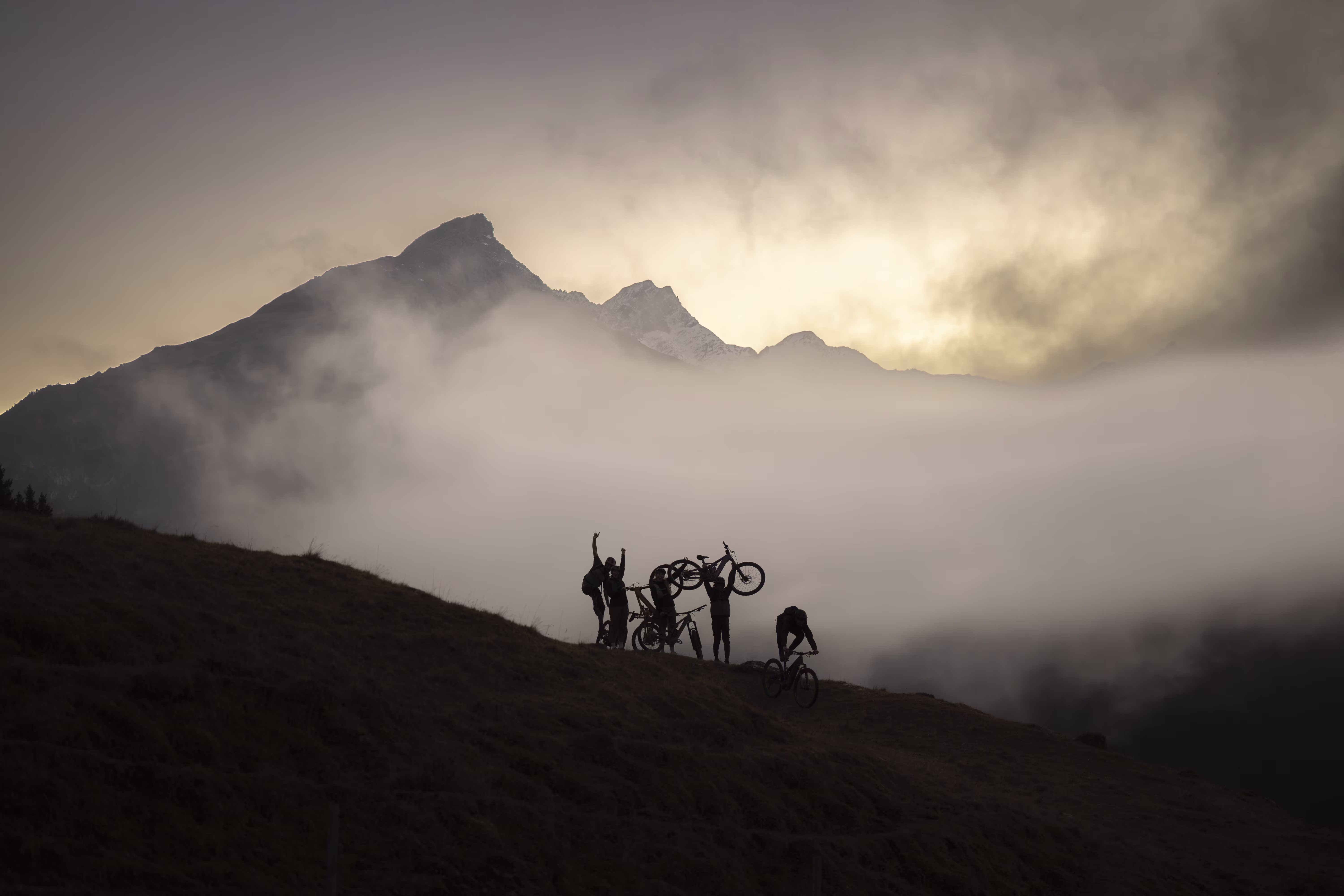 Das Bild zeigt eine Gruppe von Mountainbikern als Silhouetten auf einem Hang, umgeben von dichtem Nebel, während im Hintergrund dramatisch beleuchtete Berggipfel aus den Wolken ragen.