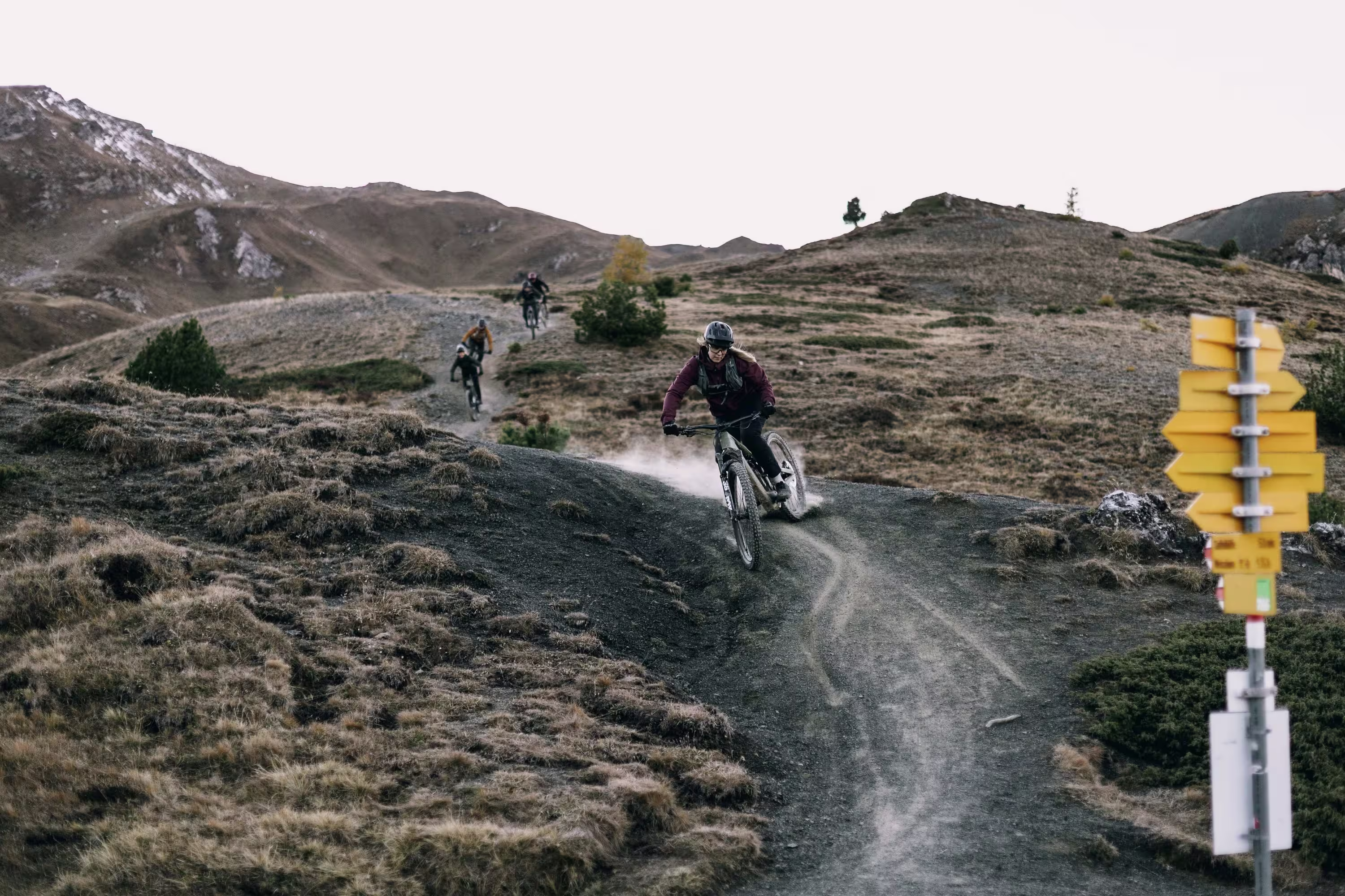 Mehrere Mountainbiker fahren in rasanter Folge über einen kurvigen Trail im hochalpinen Gelände. Der vorderste Fahrer legt sich dynamisch in die Kurve, Staub wirbelt hinter ihm auf. Rechts am Bildrand markiert ein gelber Wegweiser die Route – ein Mix aus Orientierung, Flow und Abenteuerspirit.