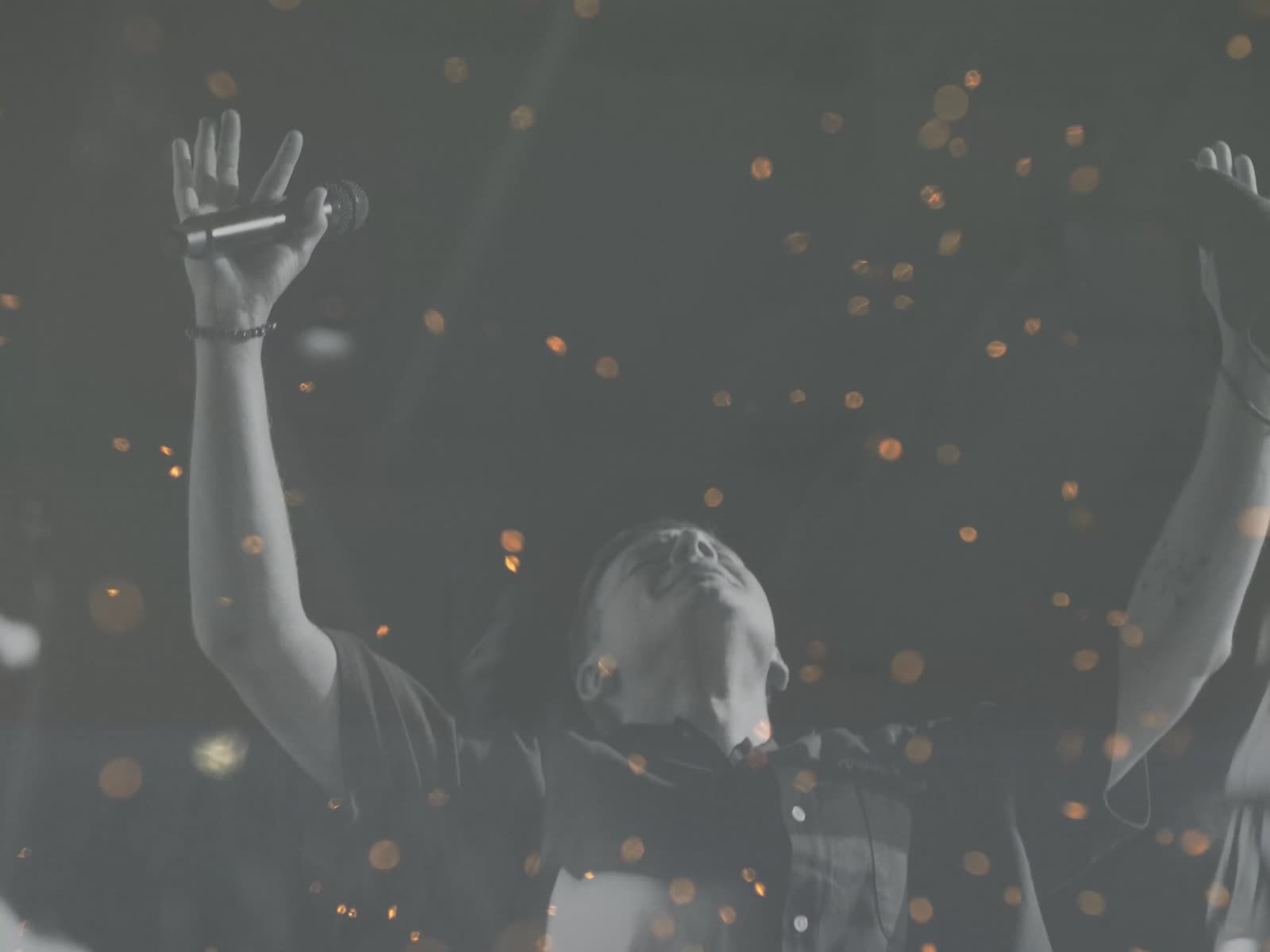 Still from the Holy Fire teaser: hands raised in worship as embers drift through the room