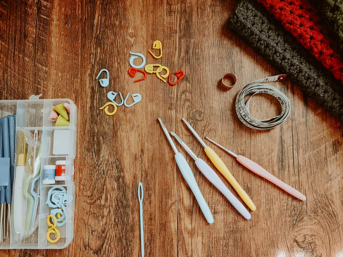 A flatlay image of crochet needles and equipment resting on a table. There is also a crocheted scarf in the corner.