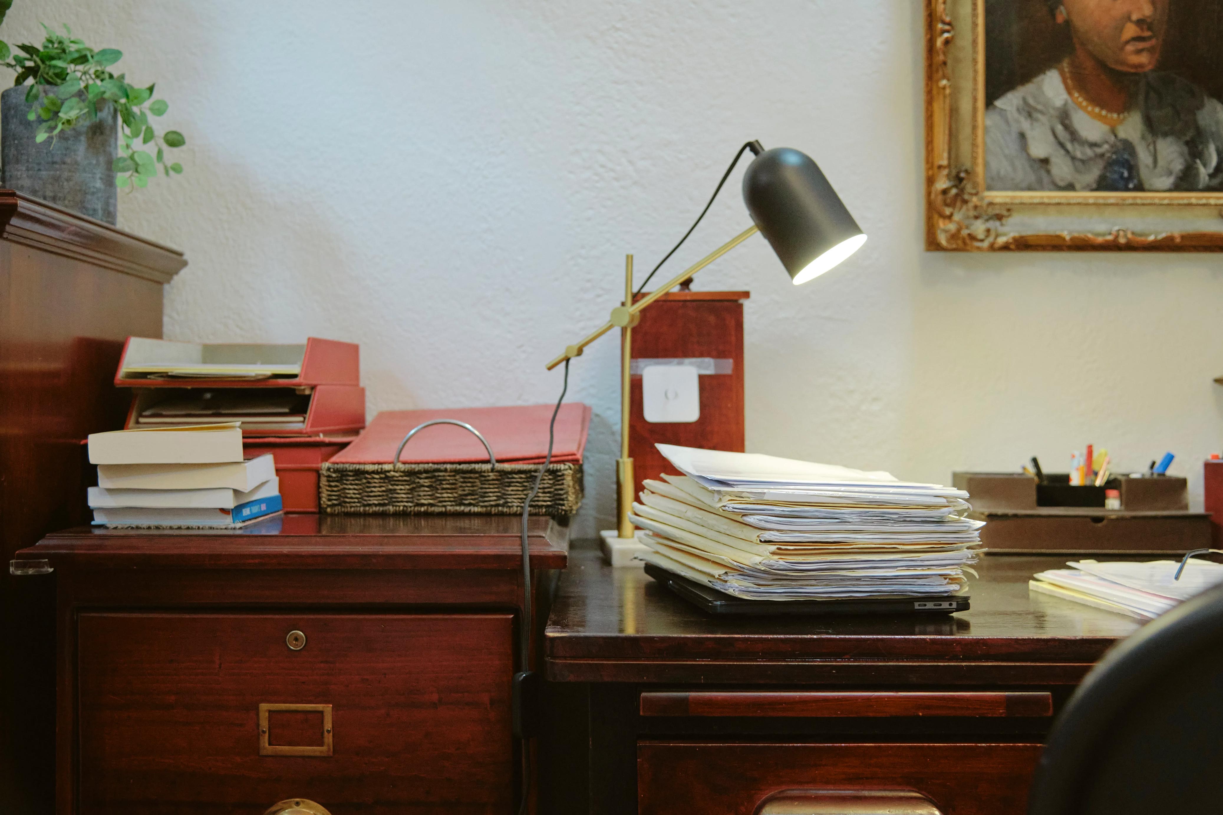 An image of a lawyer's desk. There is a lamp that is turned on, shining onto stacks of papers.