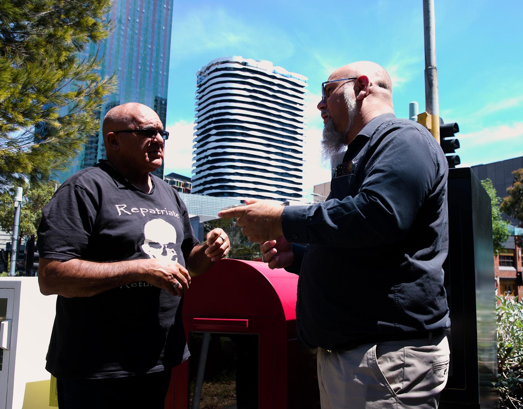 An image of two First Nations men talking. The picture is taken during daytime, and a cityscape and clear blue sky is in the background.