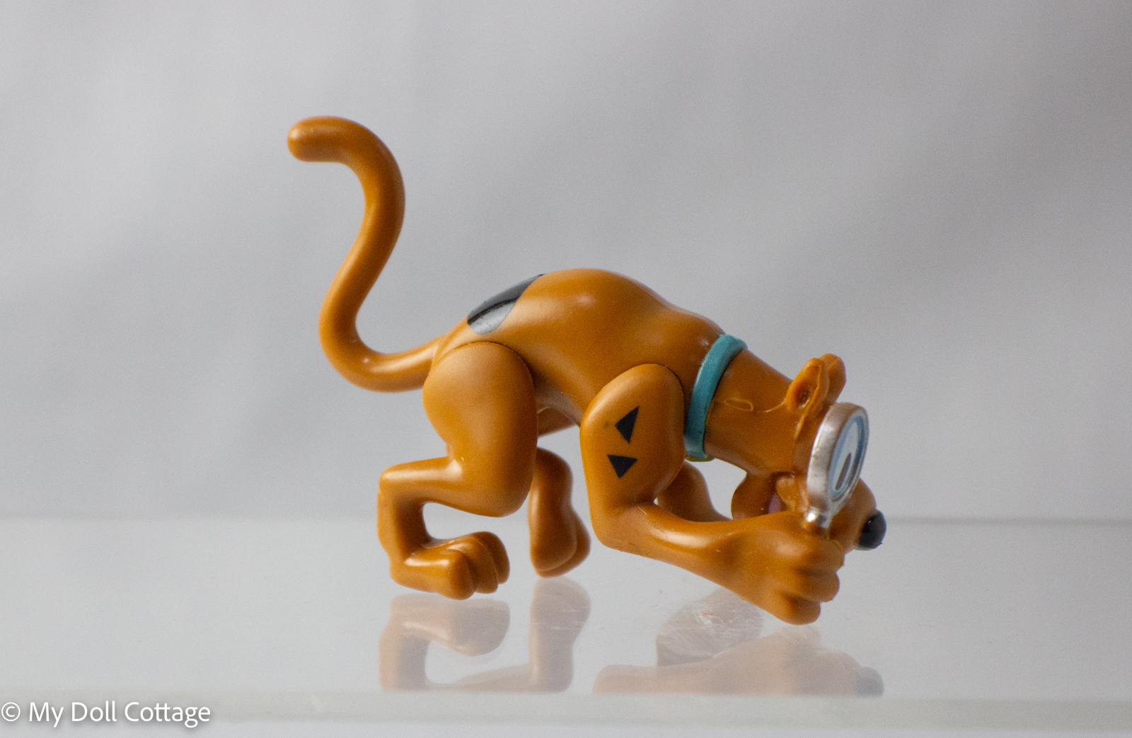 Side view of Scooby-Doo Mystery Mates figurine in detective pose, holding magnifying glass close to ground, tail curled upward.