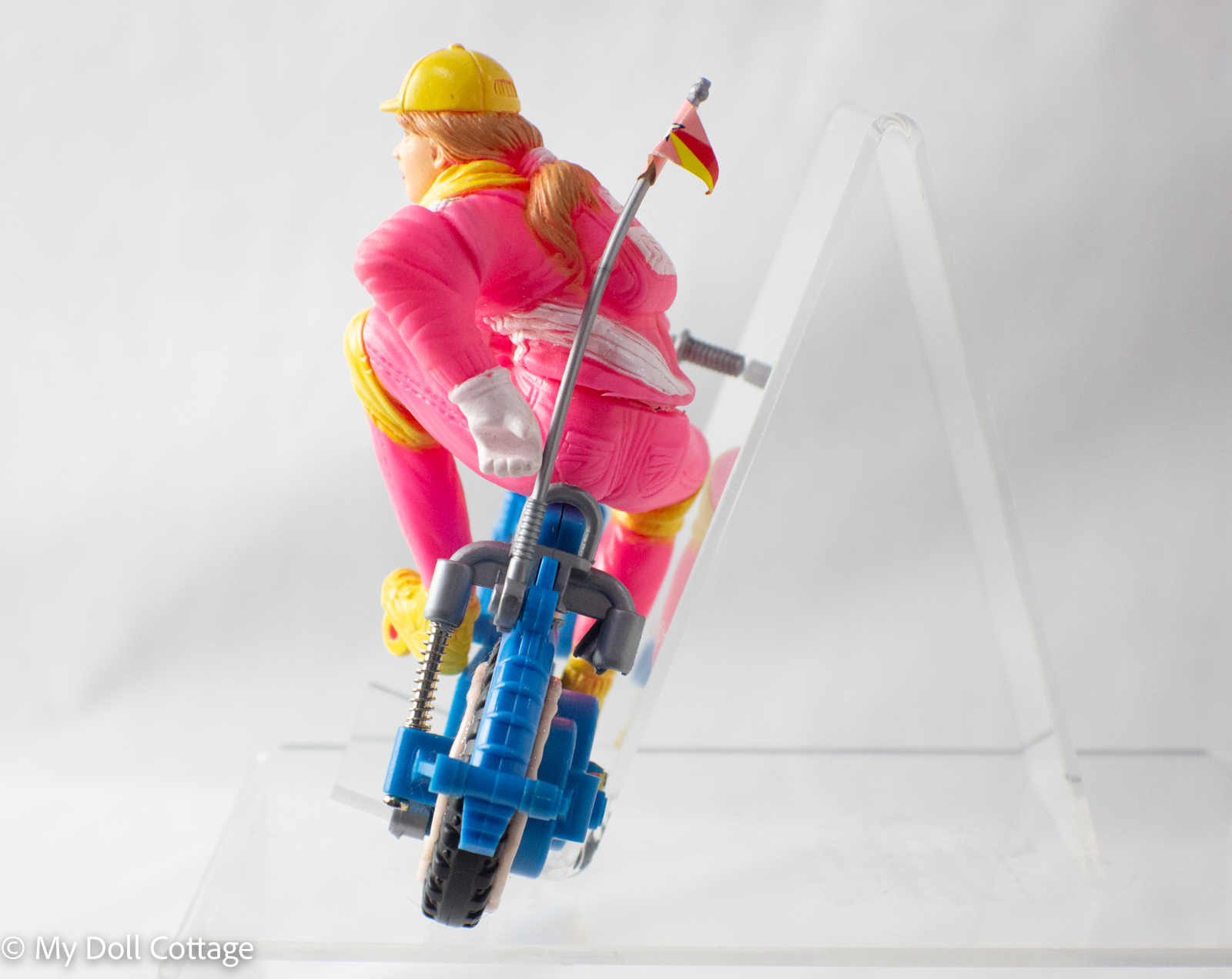 Vintage 1980s New-Ray girl in pink outfit with yellow accessories riding a blue friction-powered bicycle.