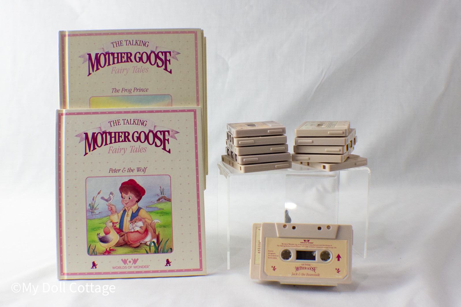 Two The Talking Mother Goose Fairy Tales books standing upright on the left, titled The Frog Prince and Peter & the Wolf. To the right, several stacks of beige cassette tapes are arranged on clear display blocks, with an additional cassette tape labeled Ja