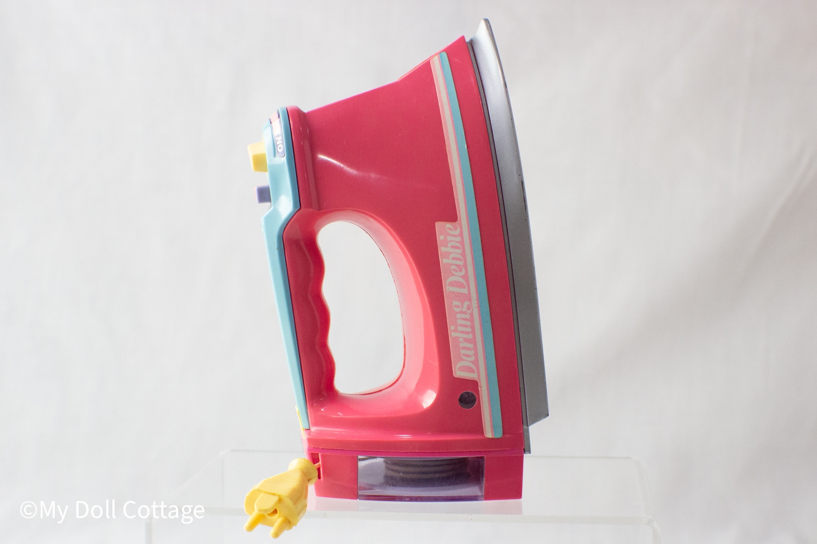 Side profile of a pink and aqua Darling Debbie toy iron with a yellow toy plug and retractable cord against a white background.