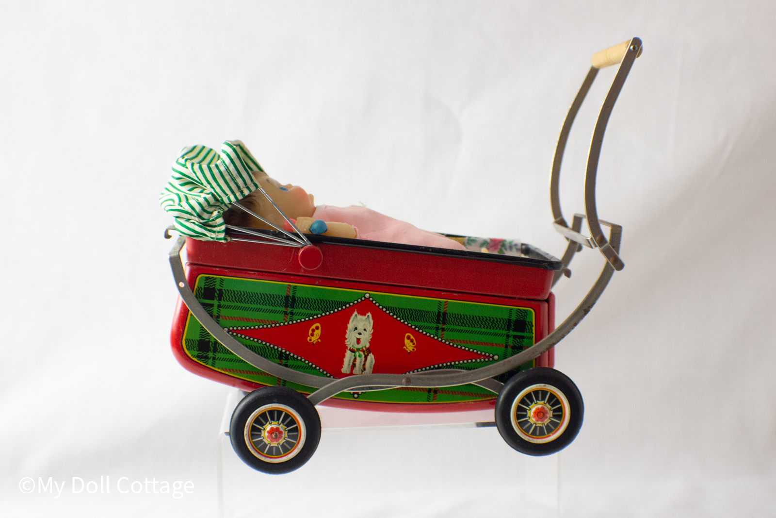 Side view of 1950s–60s Japanese lithographed tin toy pram with baby inside, red body and green plaid design.