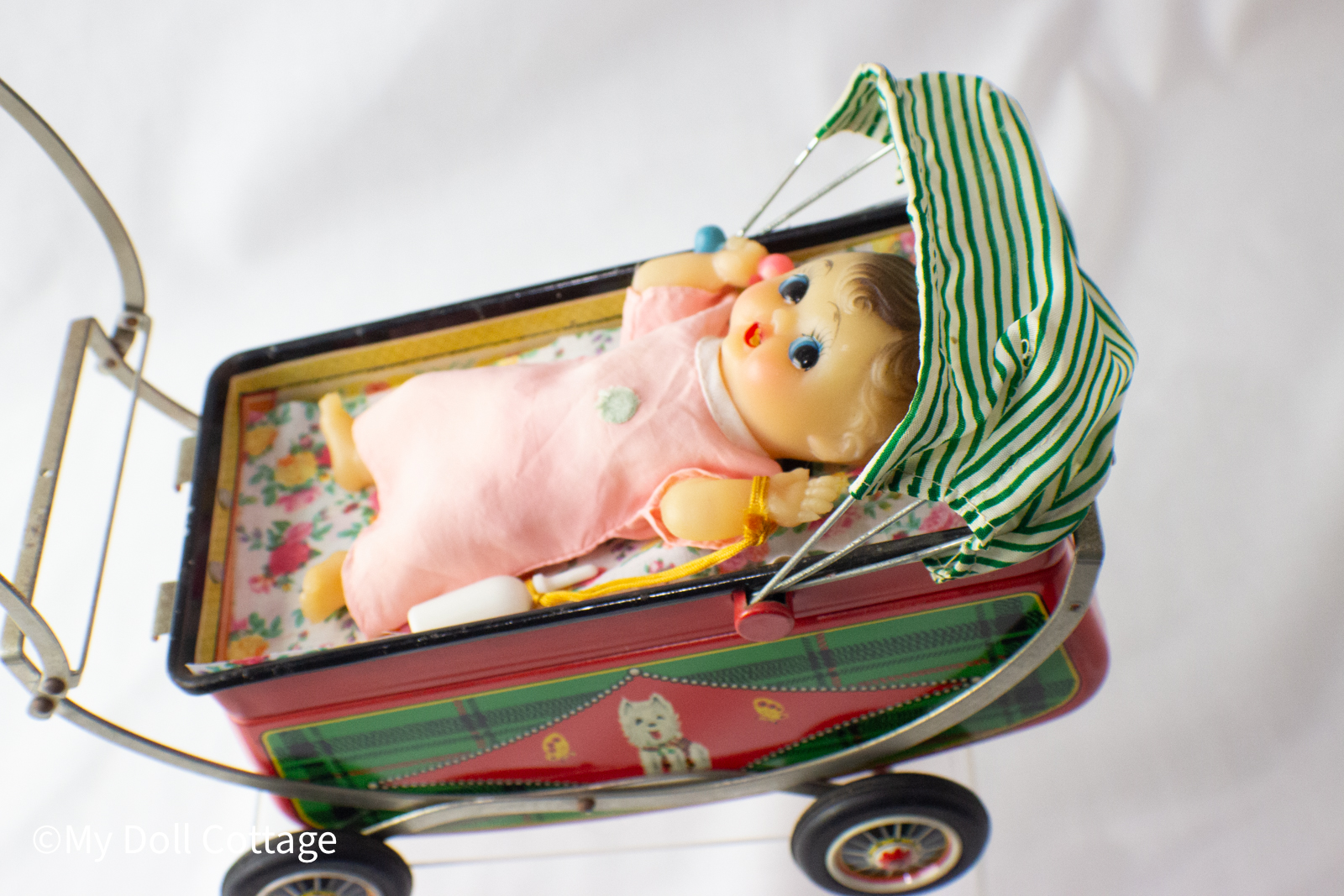 Angled view of baby doll resting in lithographed tin pram, showing canopy supports.