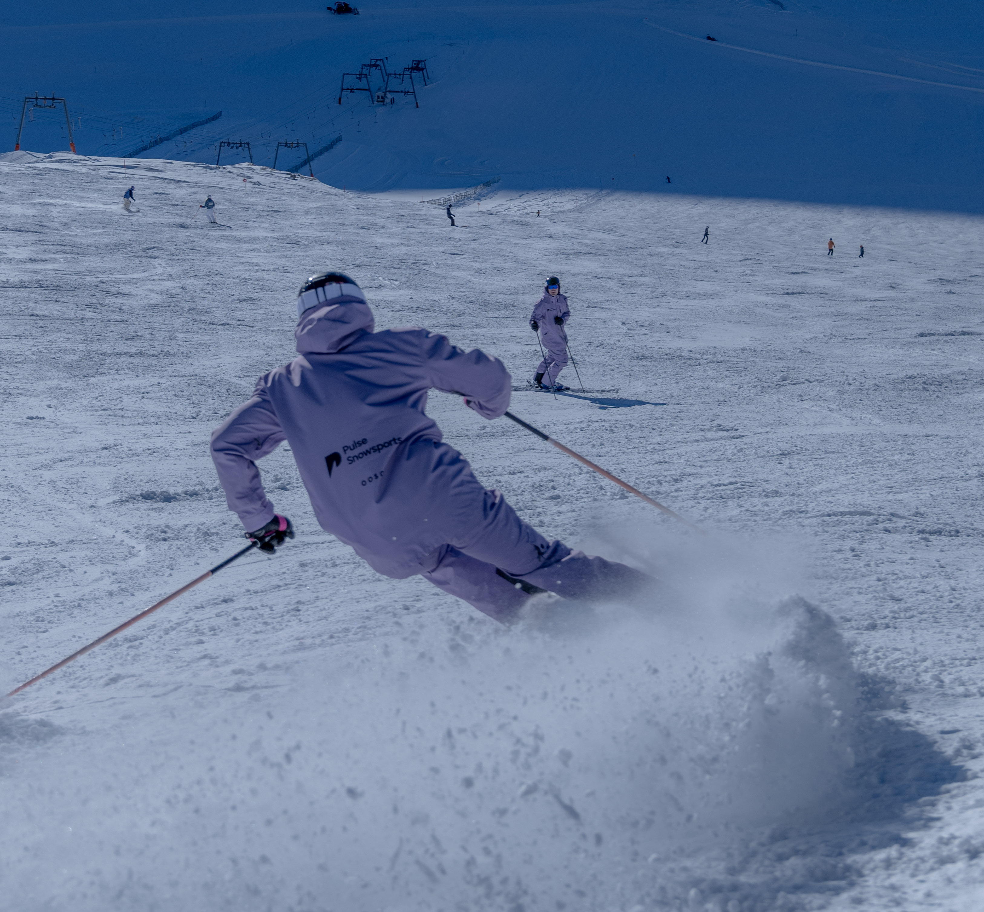 Skier in purple outfit carving a turn on a snowy slope with other skiers visible in the background.