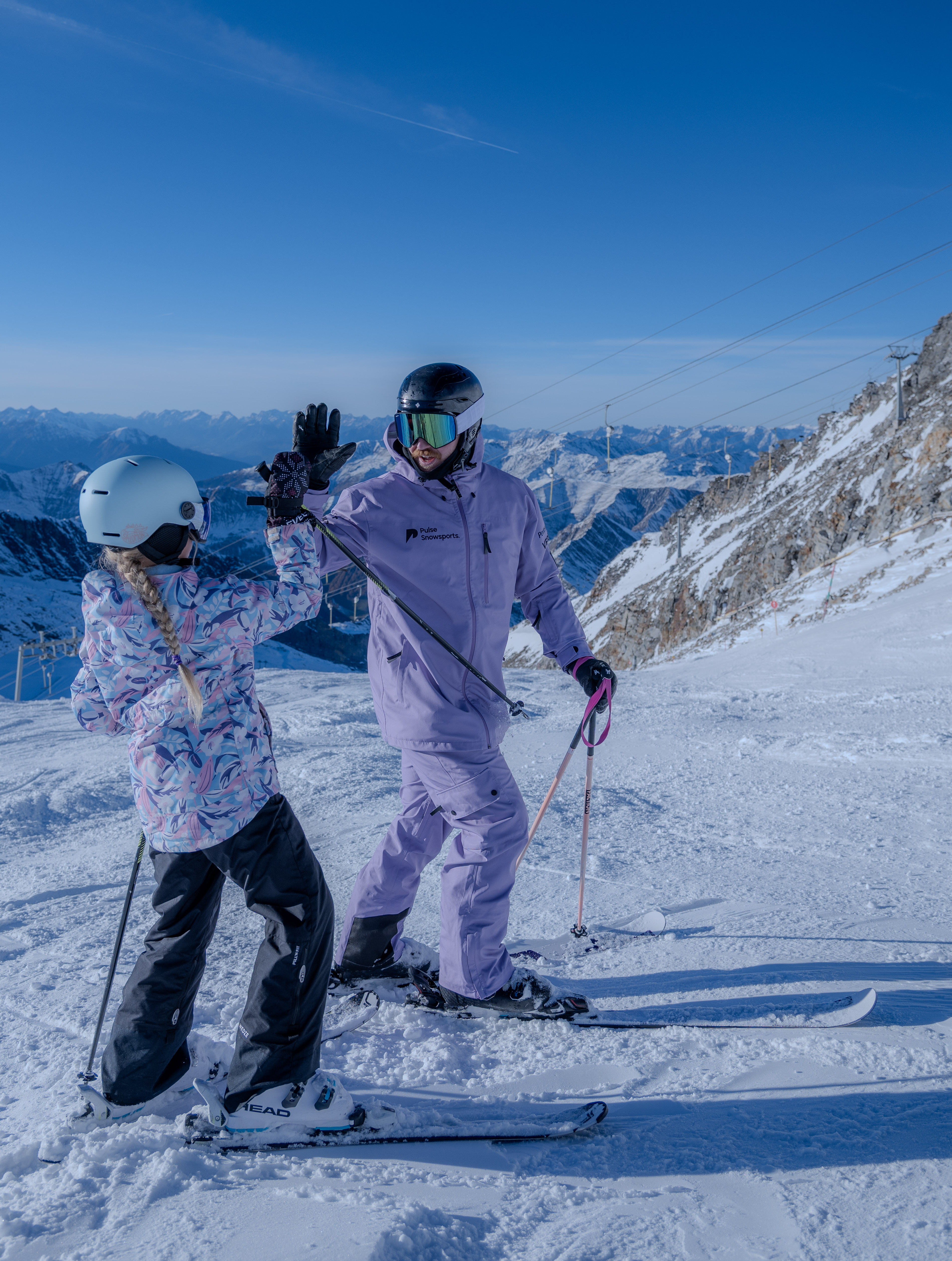Ski instructor in purple gear high-fiving a child wearing a white helmet on a snowy mountain slope with clear blue sky.