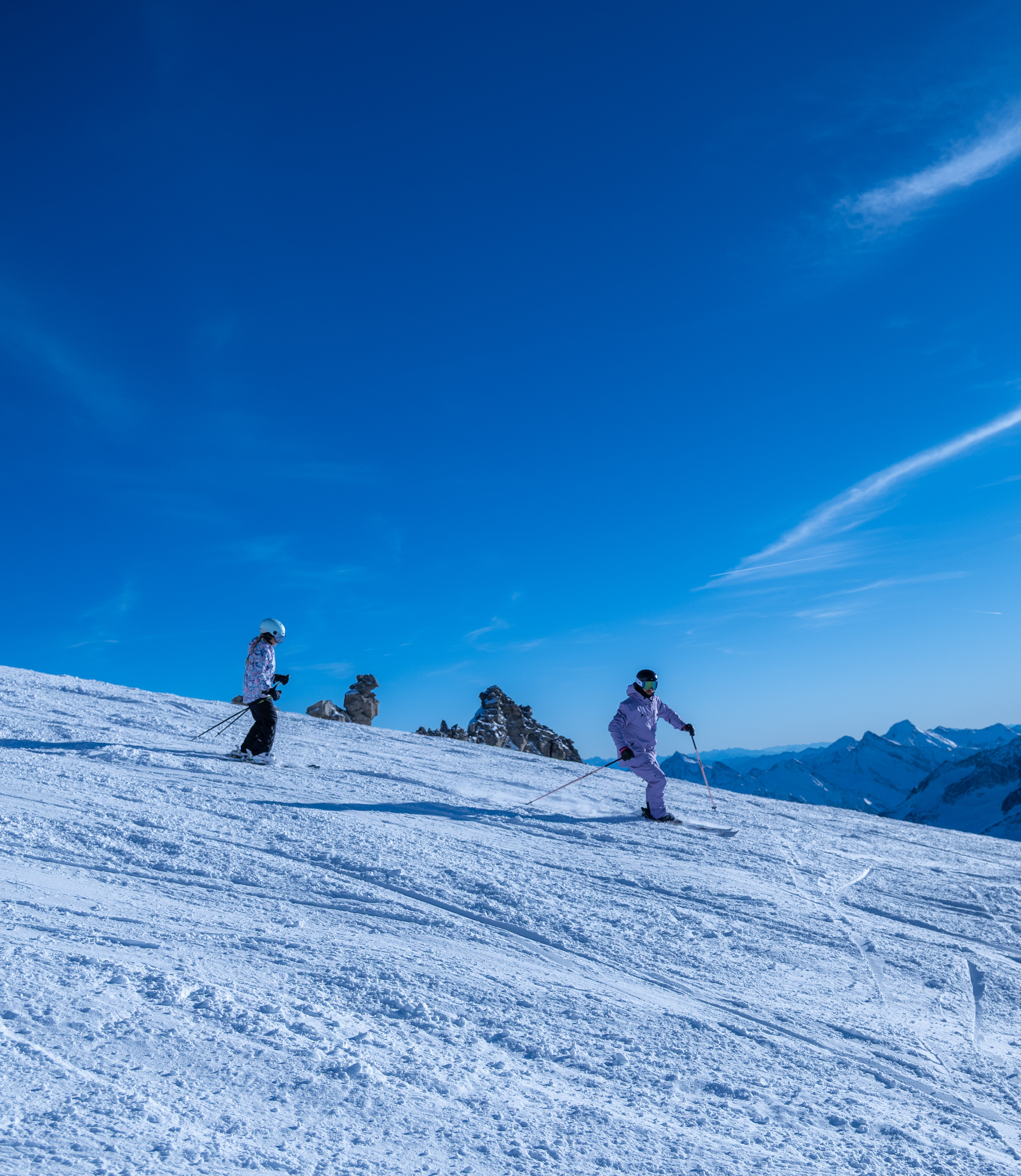 Ski instructor leading a child skiing down a snowy slope under a clear blue sky with mountains in the background.