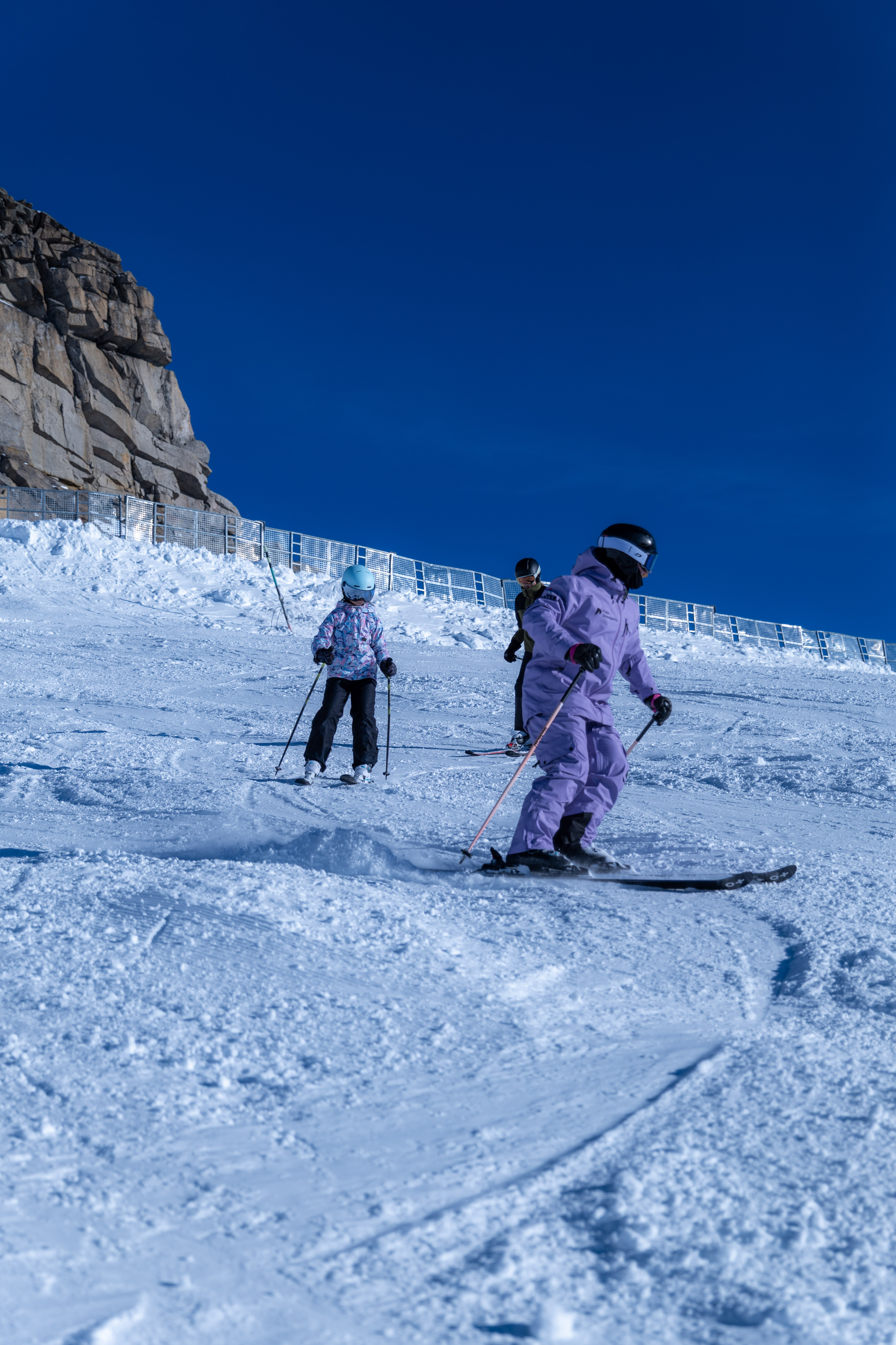 Three skiers descending a snowy slope with a rocky cliff and clear blue sky in the background.