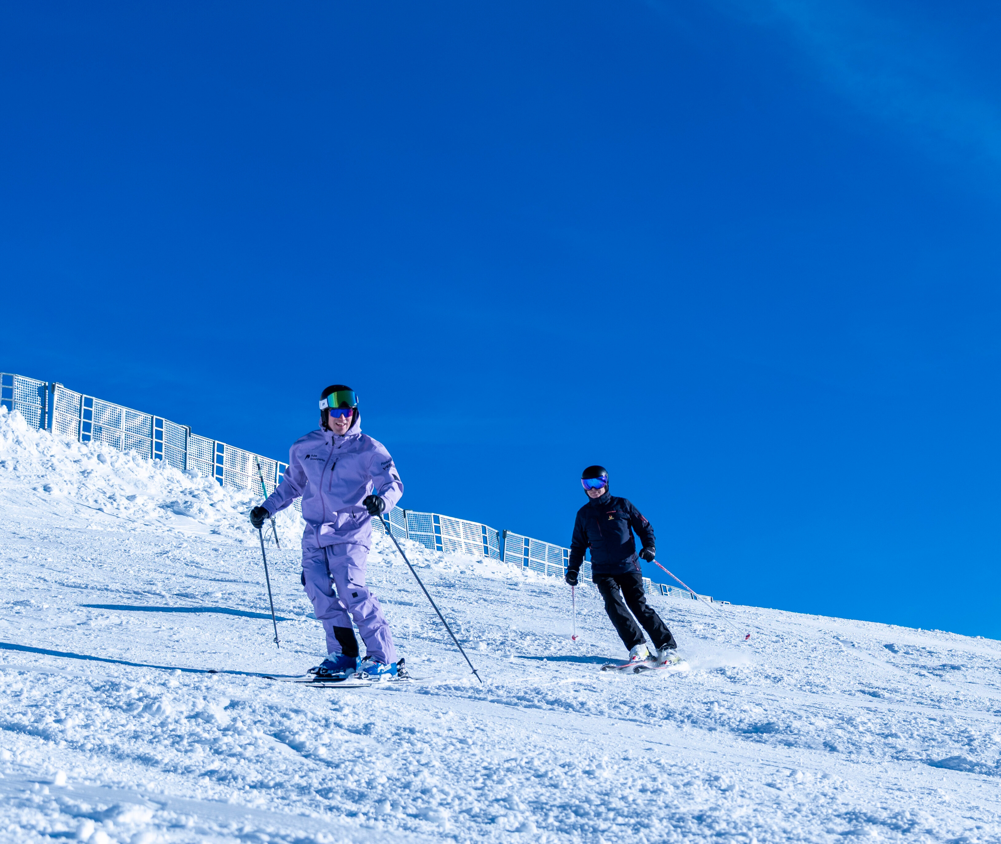 Two skiers descending a snowy slope under a clear blue sky, one wearing a lavender outfit and the other in black.