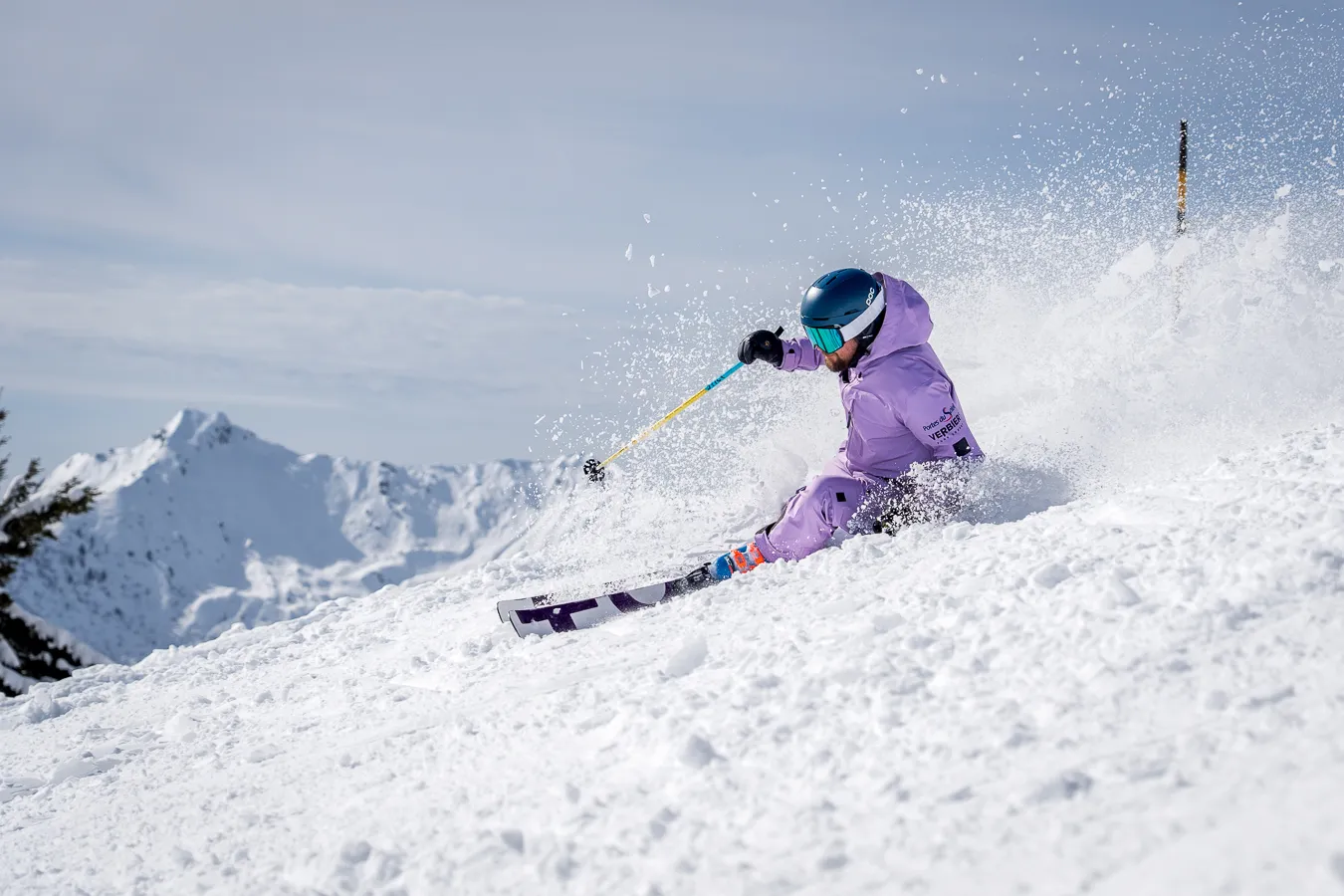 Skier in purple outfit carving through snow on a sunny mountain slope with snowy peaks in the background.
