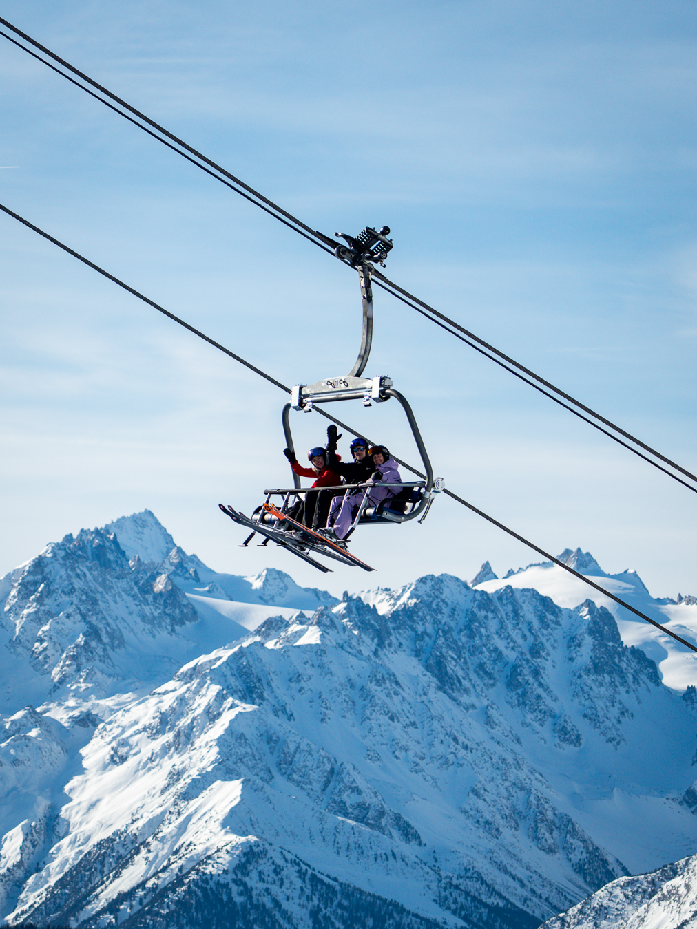 Three skiers on a chairlift over snow-covered mountain peaks under a clear blue sky.