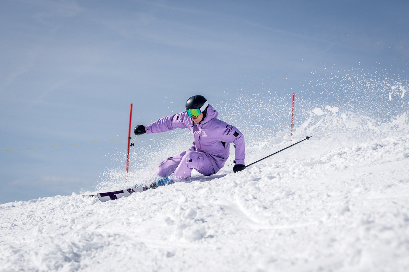 Skier in purple outfit carving a turn on a snowy slope with ski poles and goggles.
