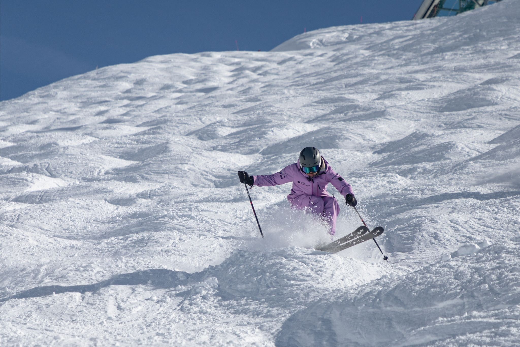 Skier in purple outfit skiing down a mogul-covered snowy slope under a clear blue sky.