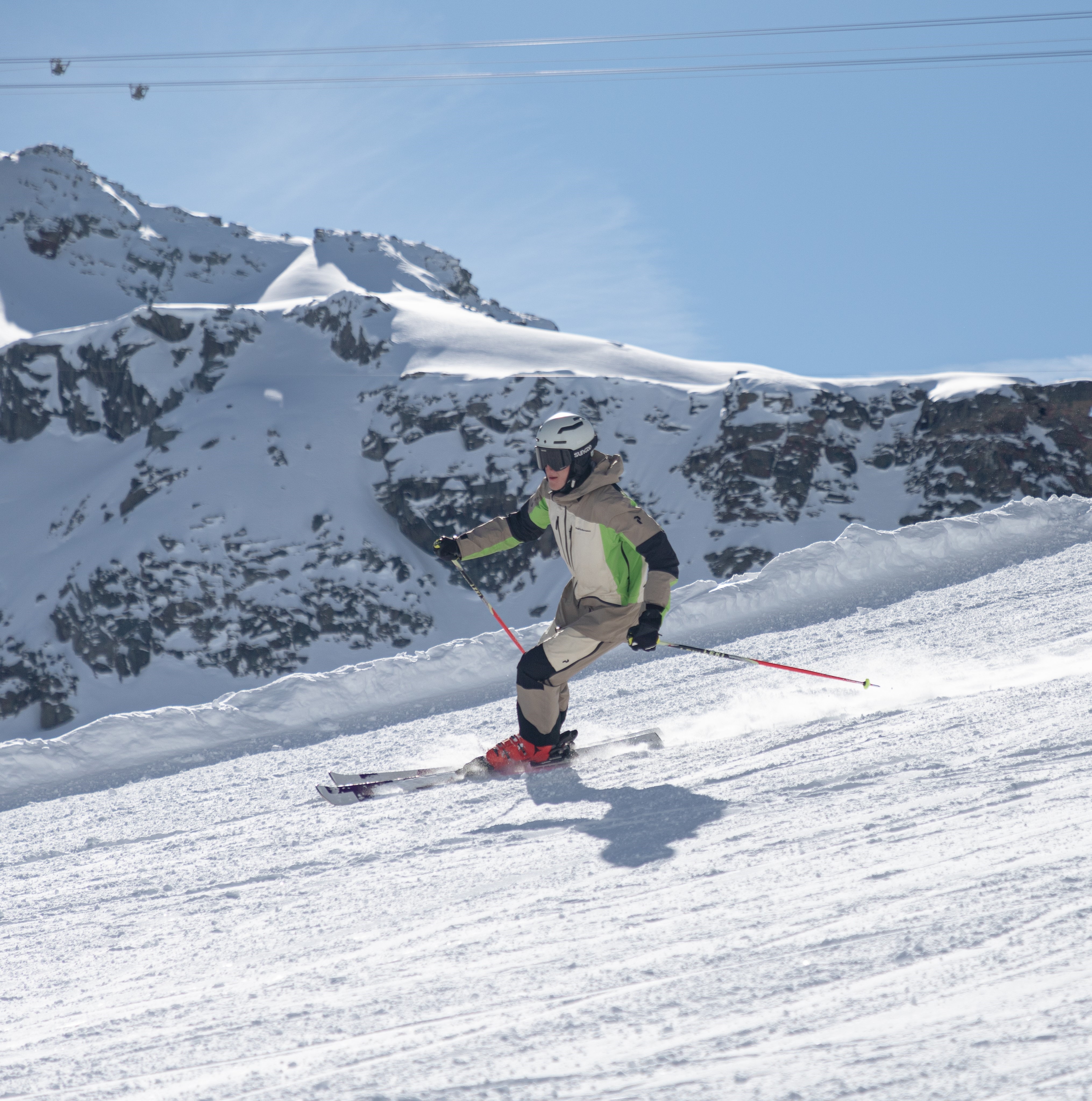 Skier in beige and green gear skiing down a snow-covered slope with snowy mountains in the background.