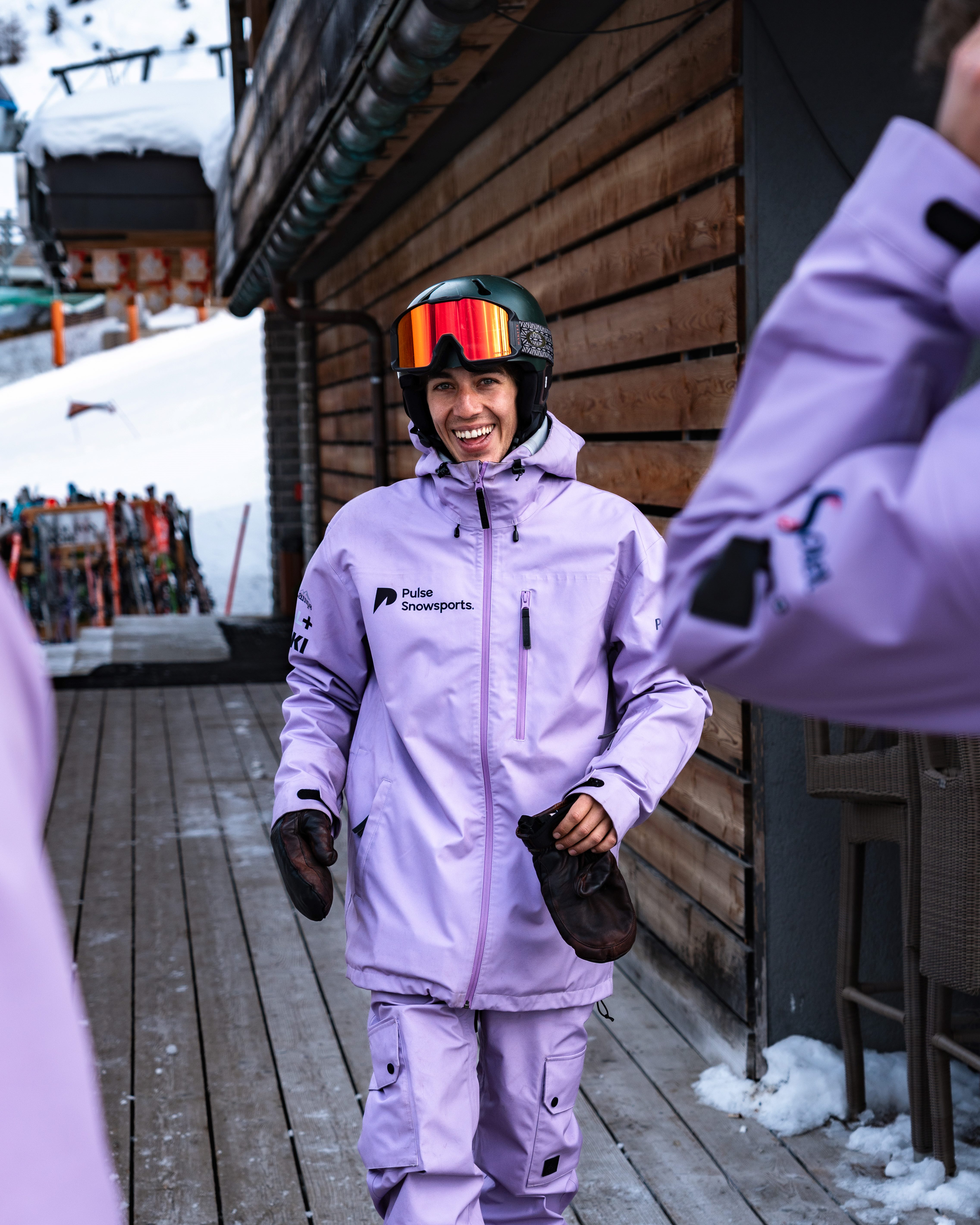 Smiling skier in purple jacket and pants holding mittens, standing on wooden deck near snow-covered slope.