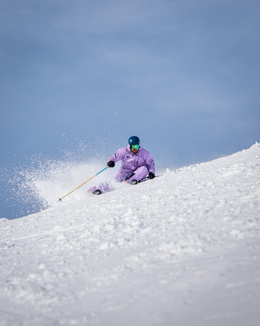 Cameron Skinner, Pulse Snowsports ski instructor, performing an hip-down carve towards the camera, spraying snow from his skis against a backdrop of clear blue skies.