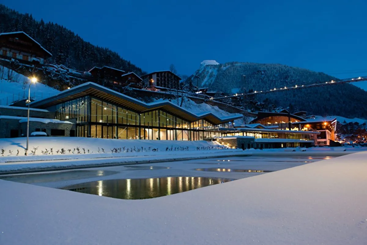 Morzine Aquatic centre lit up by lights at night with soft snow around the outdoor swimming pool and the Morzine suspension bridge in the backround