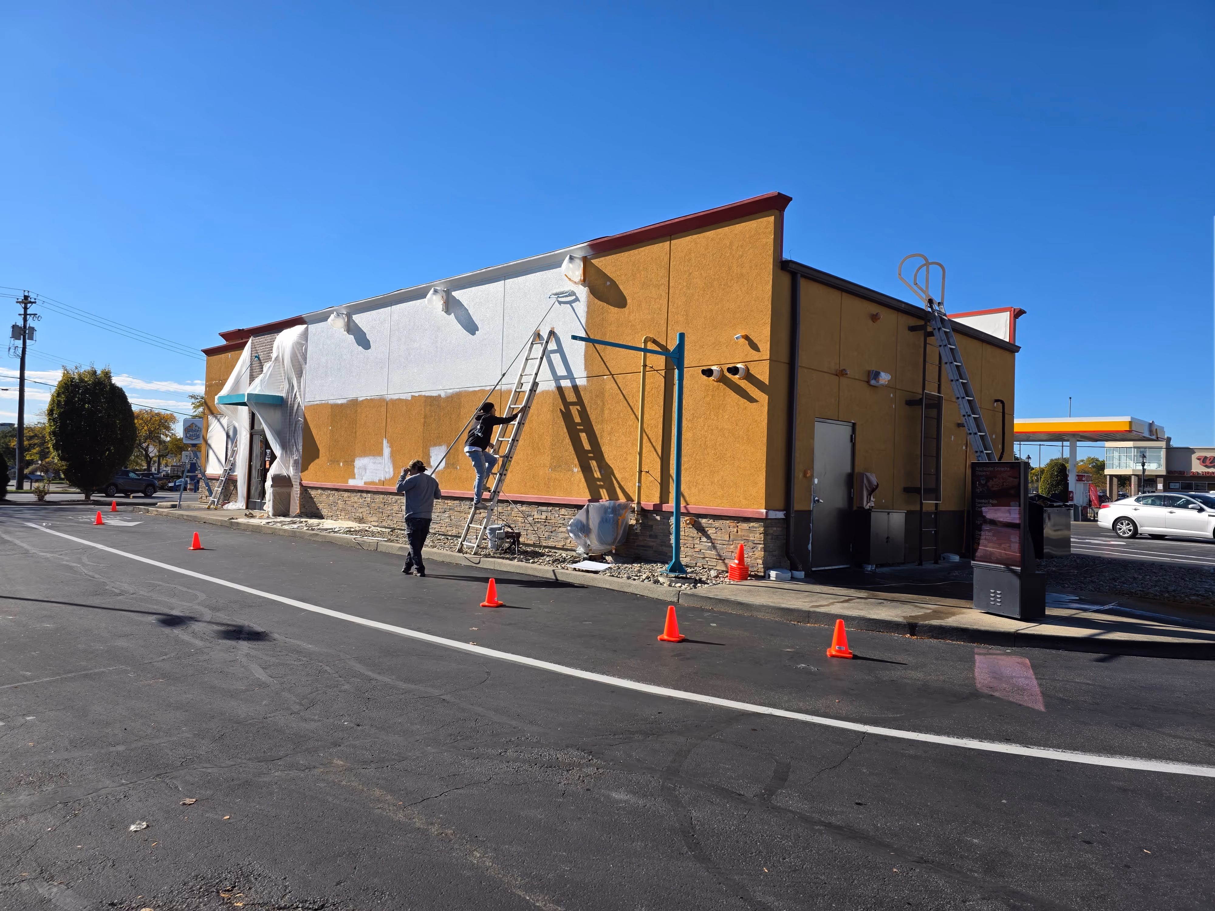Workers painting the exterior wall of a restaurant building with ladders and orange safety cones placed along the pavement.