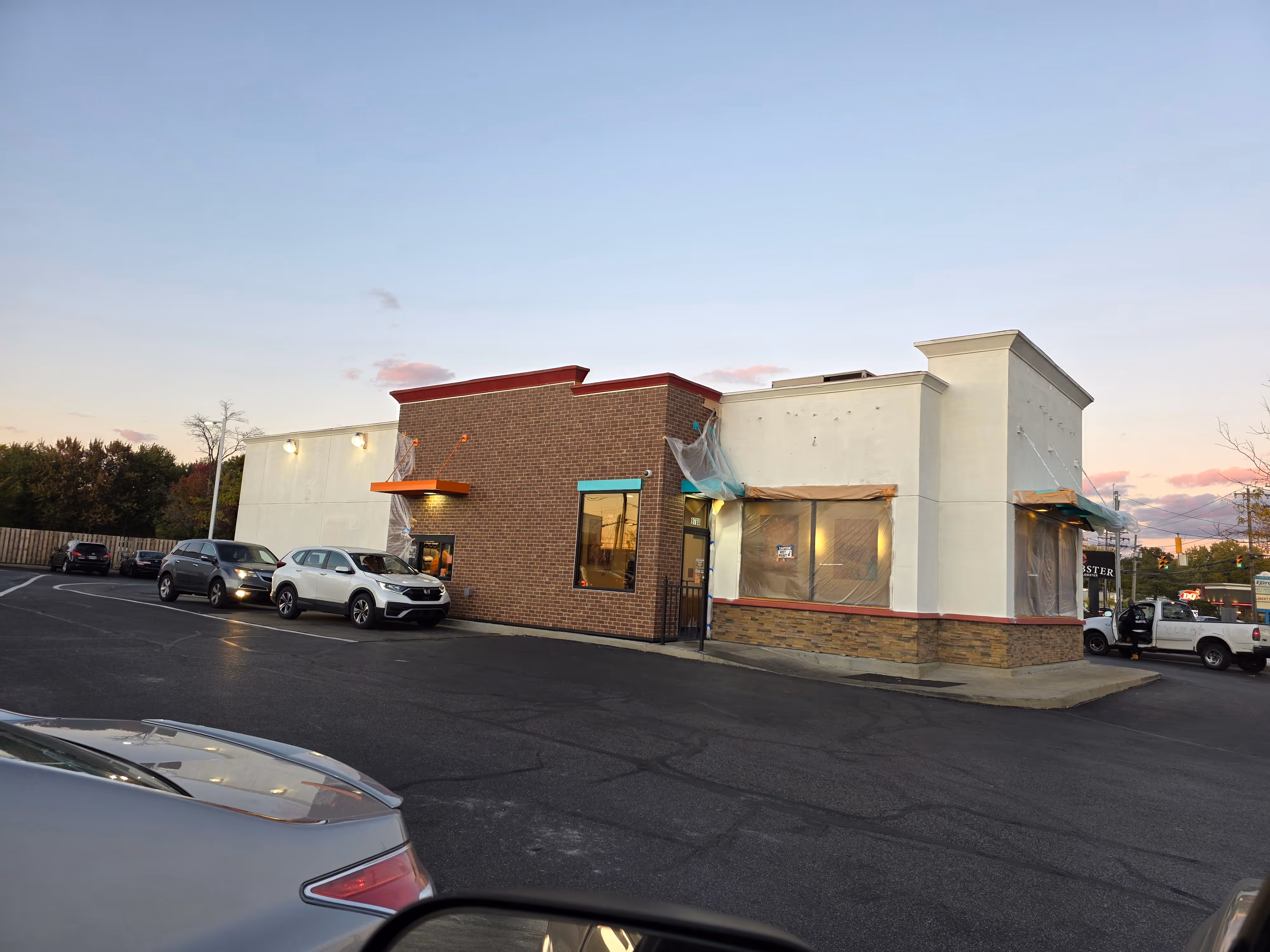 A fast food restaurant with brick and white walls undergoing renovation, cars parked in front during dusk.