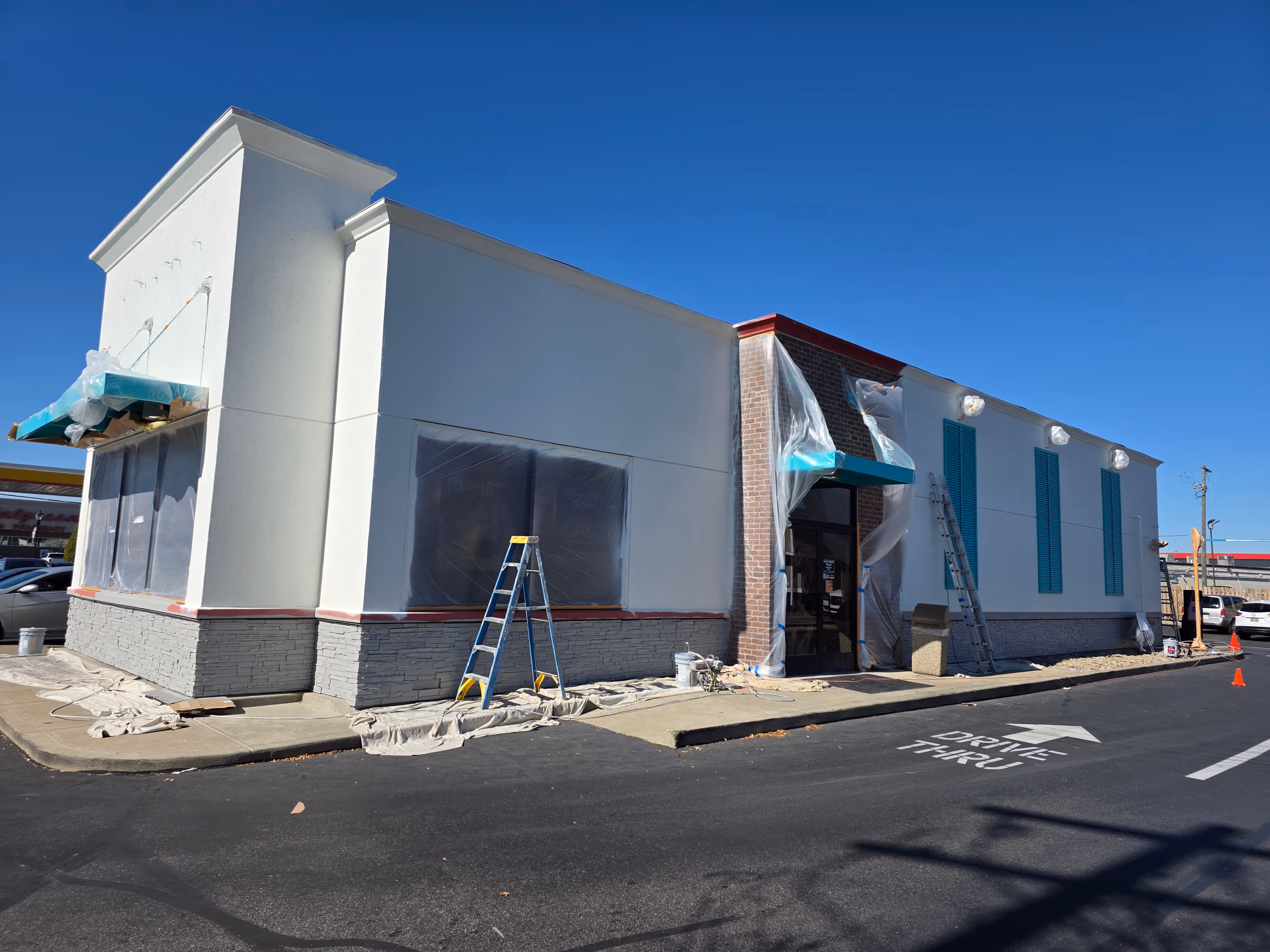 Building under renovation with white and gray exterior, ladders, covered windows, and a drive-thru lane on a sunny day.