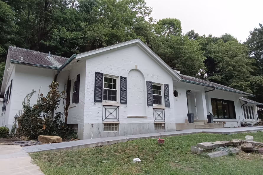 White brick house with dark shutters surrounded by green trees and a grassy yard.