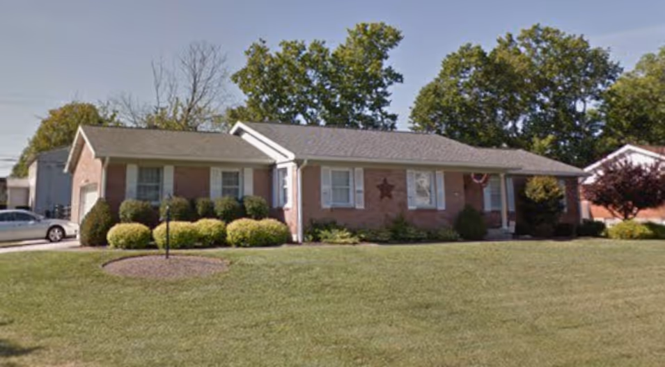 Single-story brick house with a gray roof, white shuttered windows, manicured bushes, and a large green lawn in front.