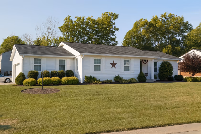 Single-story white house with dark roof, front yard shrubs, a large star decoration on the wall, and trees in the background.