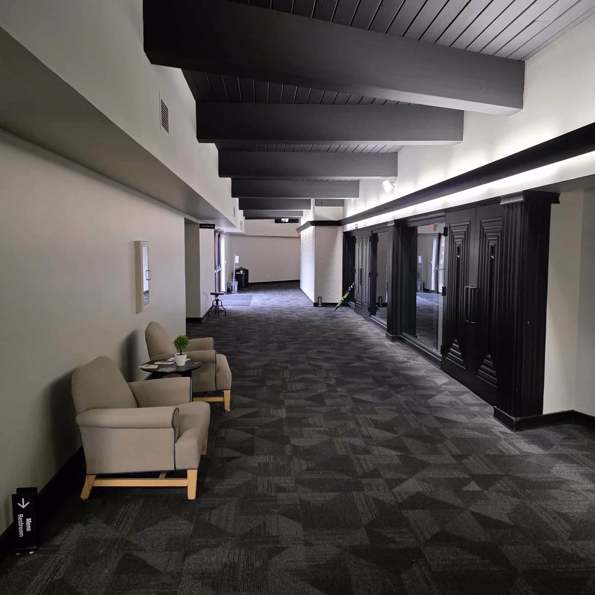 Empty hallway with two beige armchairs and a small table on the left, dark gray patterned carpet, black ceiling beams, and black framed glass doors on the right.