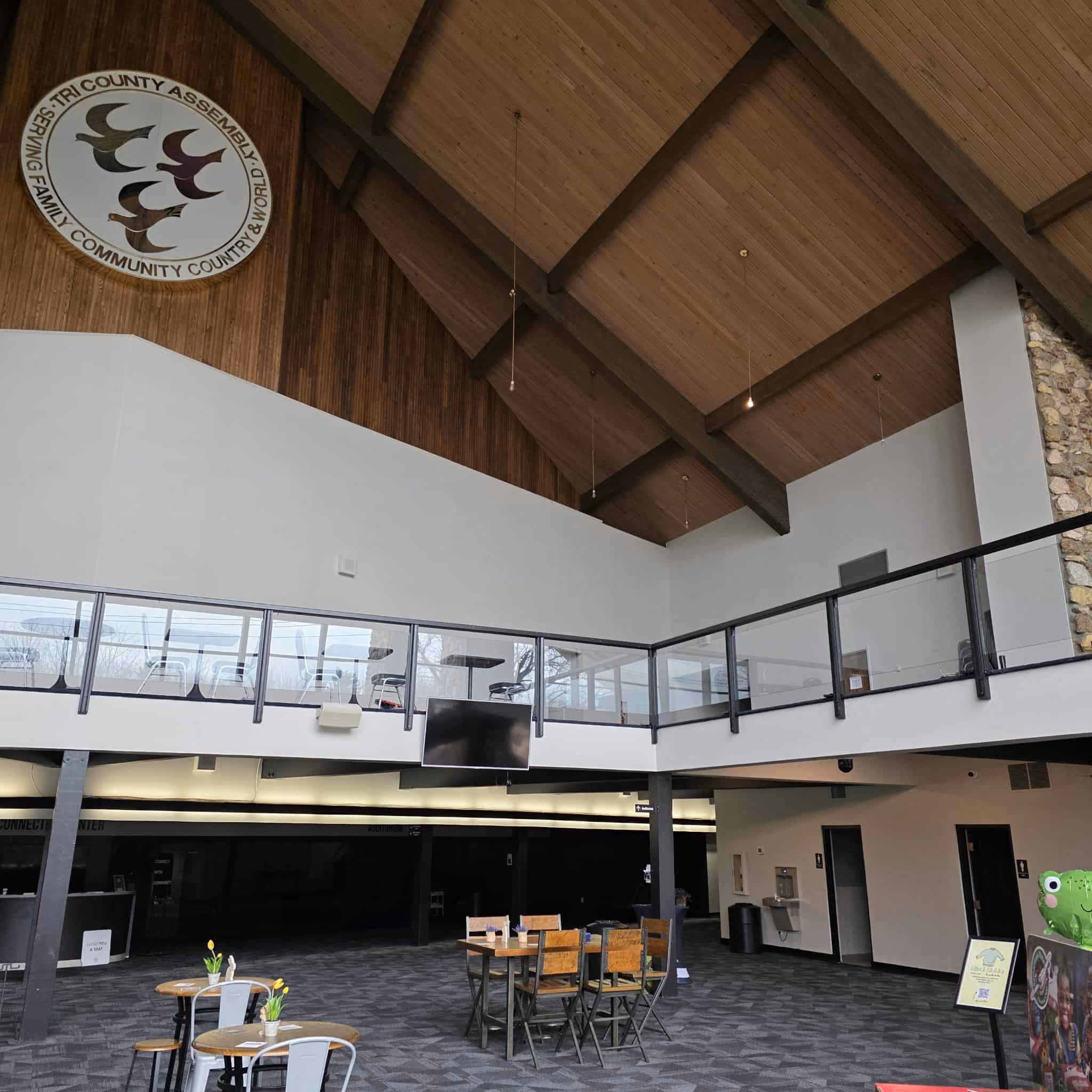 Spacious interior of a community center with high wooden ceiling, seating area with tables and chairs, and a round logo on the wall reading 'Tri County Assembly Serving Family Community Country & World'.
