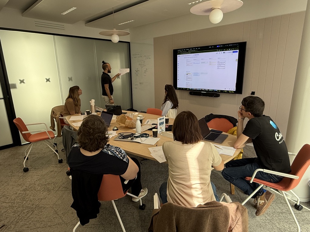 Groupe de six personnes en réunion dans une salle de conférence, avec un homme debout expliquant un diagramme sur un grand écran mural.