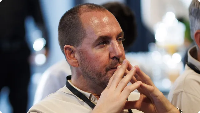 Man met kort haar en baard zit bedachtzaam met de vingers tegen de mond in een wazige binnenomgeving.