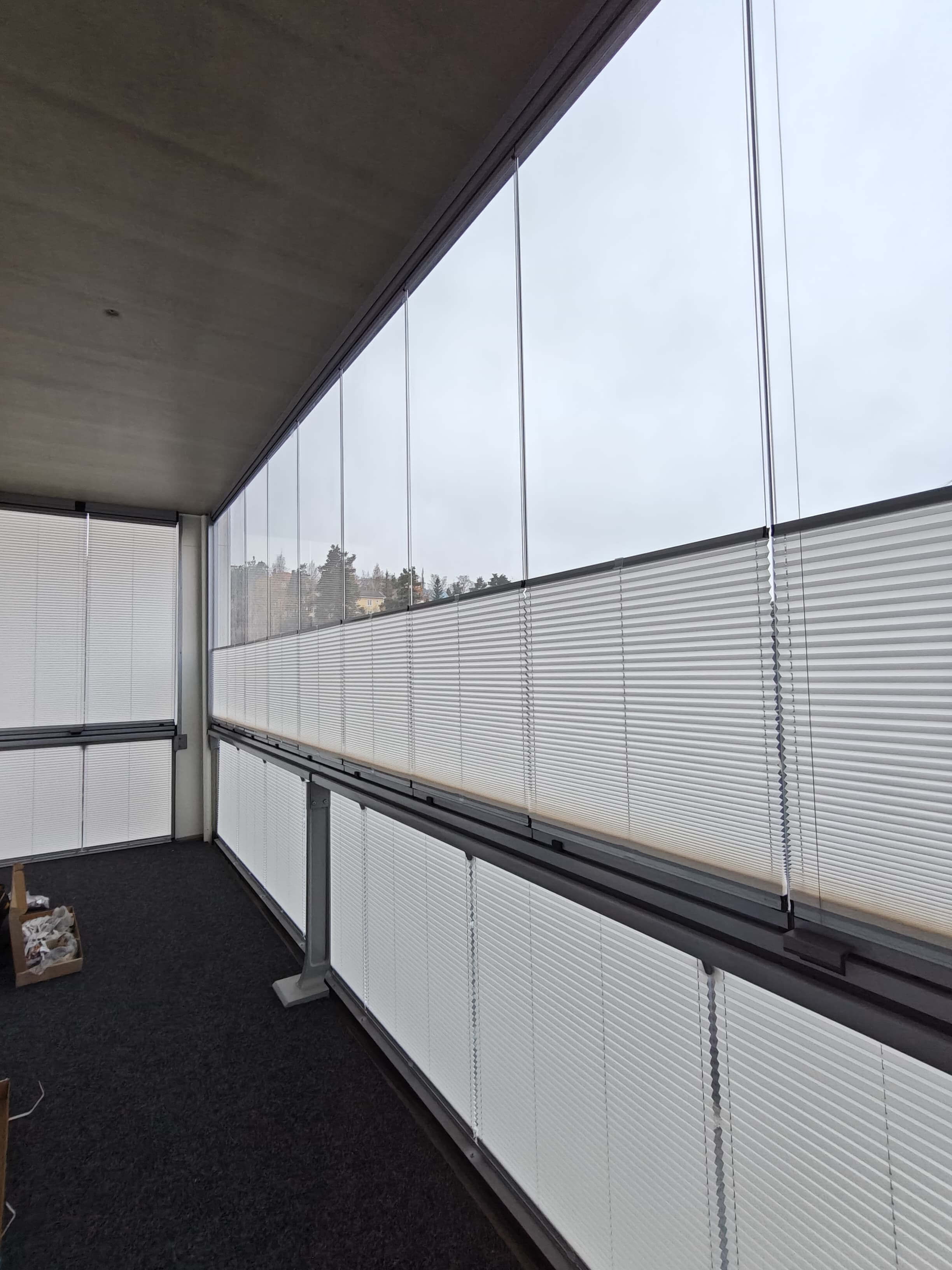 Lasitettu parveke, jossa valkoiset sälekaihtimet puolivälissä ikkunoita ja tummanharmaa matto lattialla.