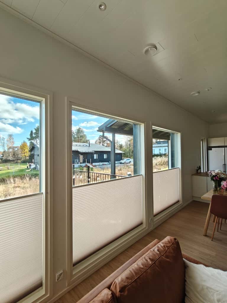 Olohuoneen sisätiloissa näkyy kolme suurta ikkunaa, joissa puoliläpinäkyvät kaihtimet, ruskea sohva, ruokapöytä kukkakimpulla ja näkymä ulos pihalle ja naapuritalolle.