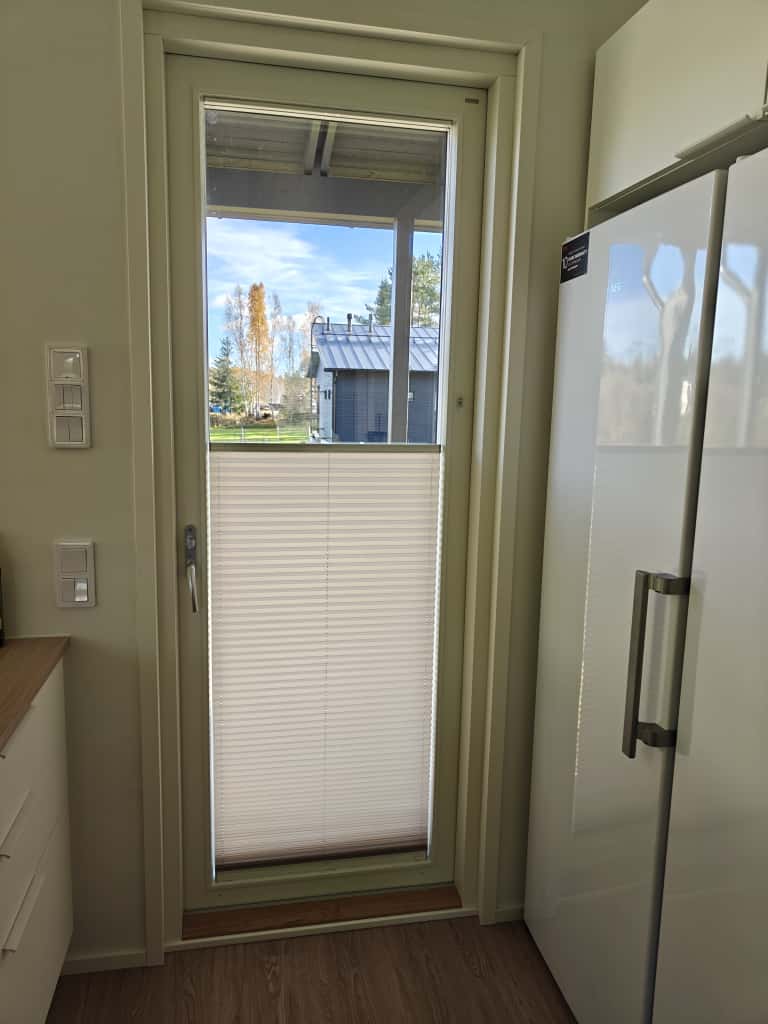 Keittiön lasinen ulko-ovi, jossa puoliksi vedetyt valkoiset sälekaihtimet, vieressä valkoinen jääkaappi ja puulattia.