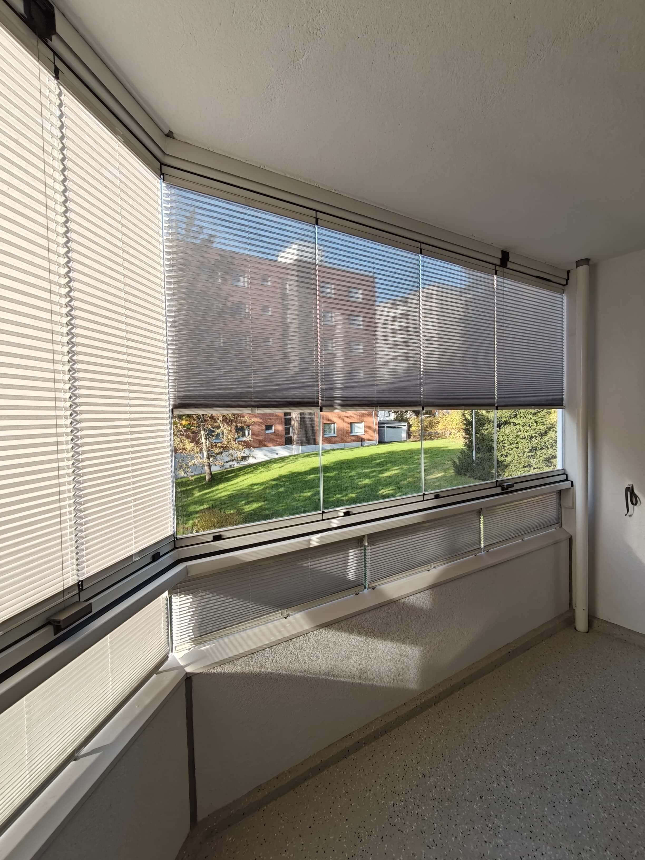 Lasitettu parveke, jossa on sälekaihtimet puoliksi alhaalla, näkyvissä viereisen rakennuksen punatiilinen julkisivu ja nurmikko.