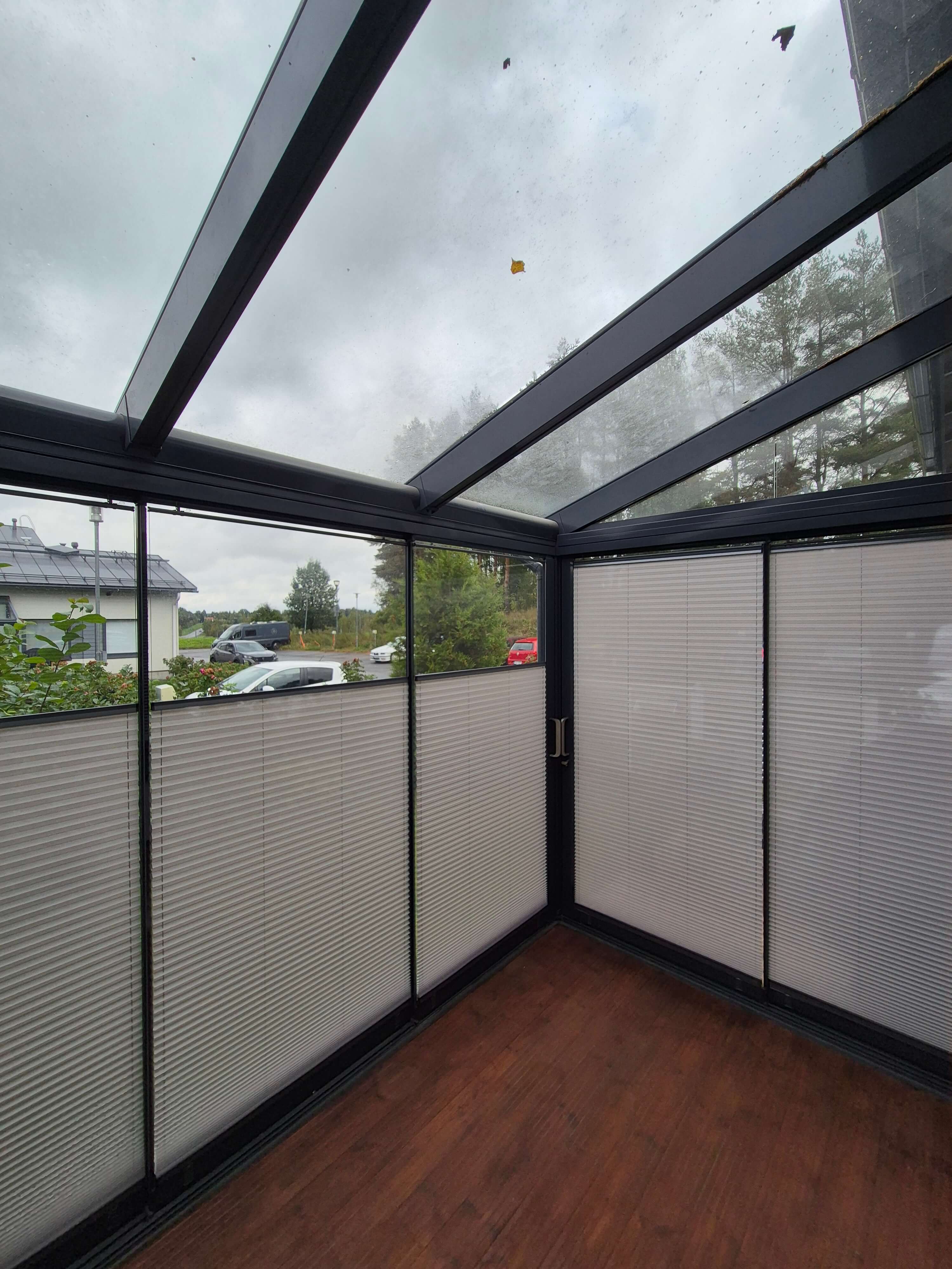 Lasitettu terassi, jossa puoliksi vedetyt pimennysverhot ja puulattia, ulkona pilvinen sää ja puita.