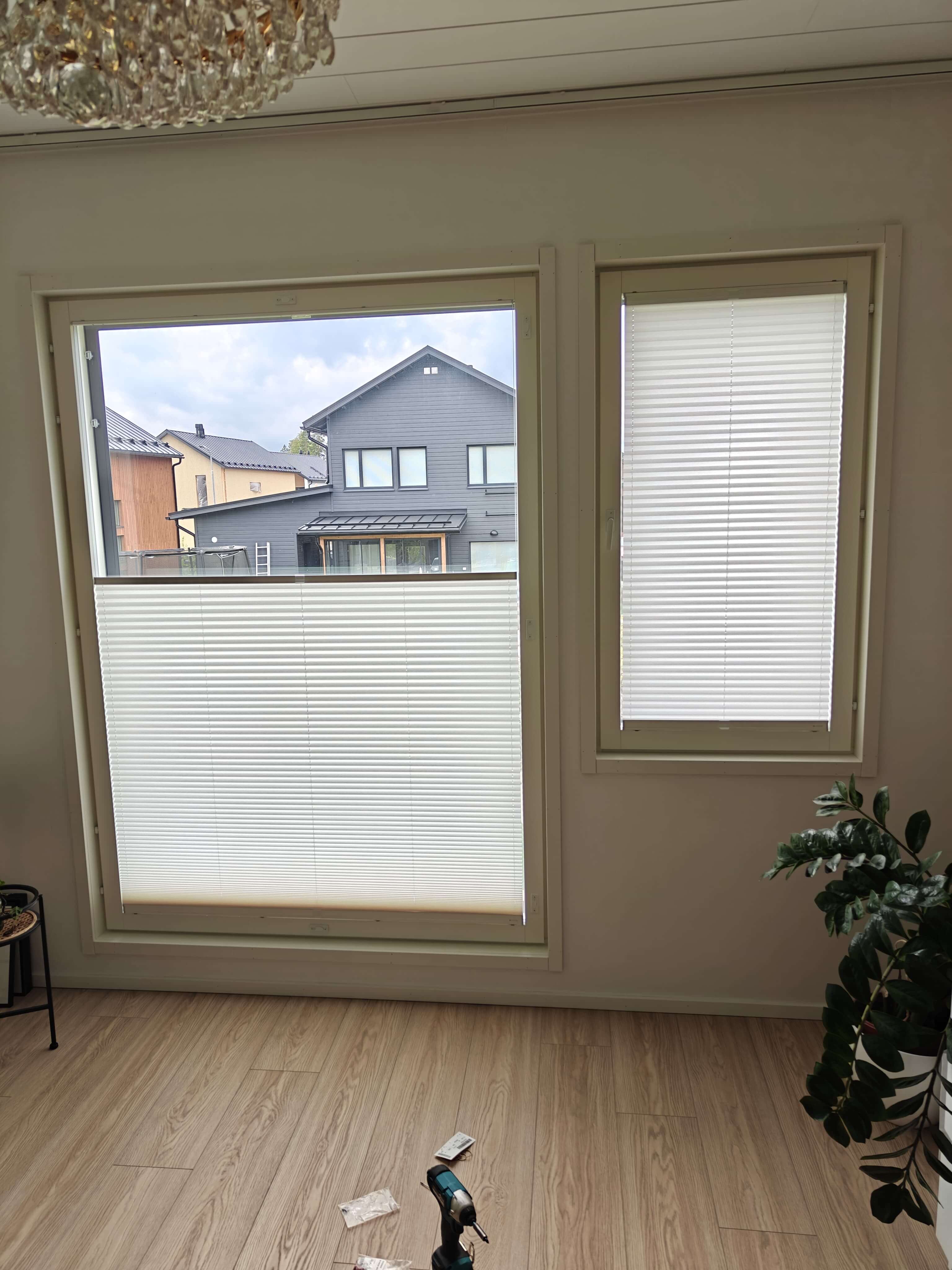 Sisäkuva huoneesta, jossa kaksi valkoista kaihdinta, avoin ikkuna näyttää harmaan talon ja puulattian, lattialla leikkuri ja kasvi oikealla.