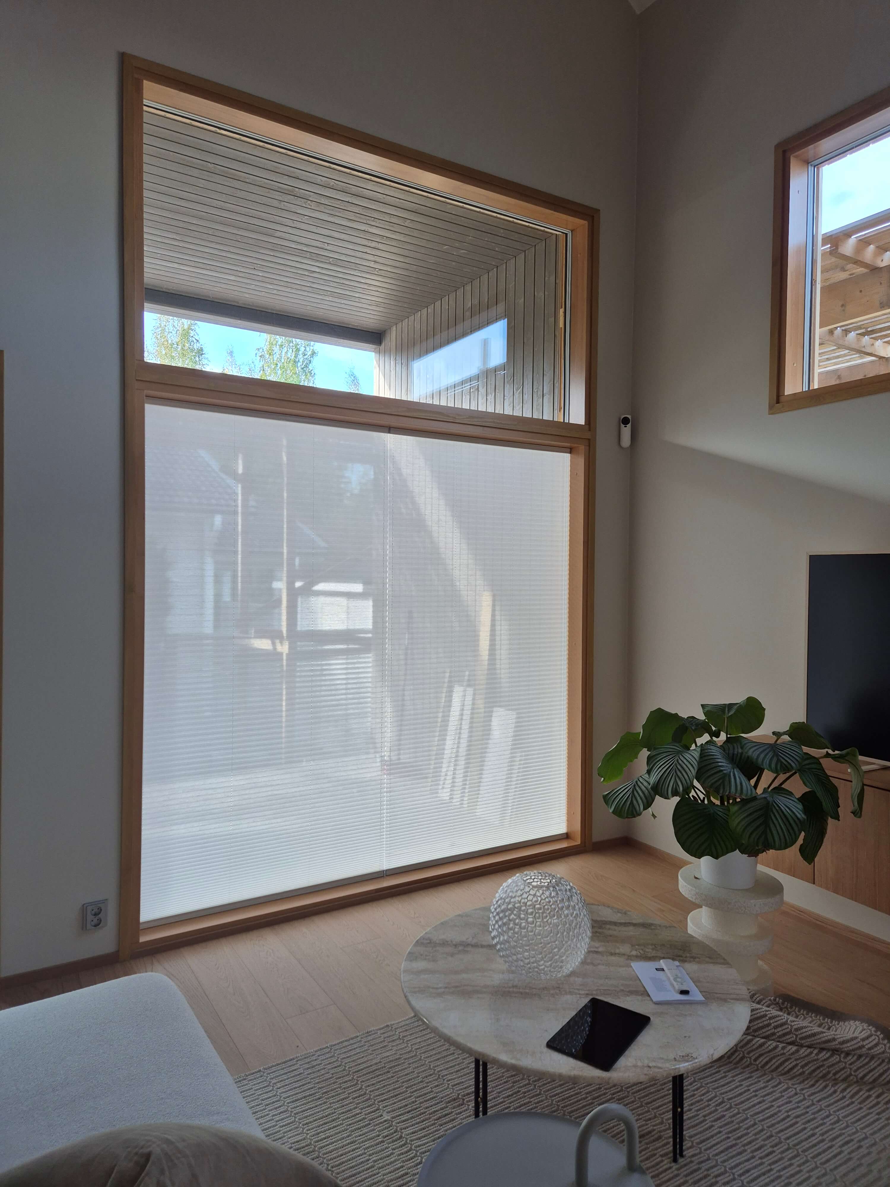 Moderni olohuone, jossa suuri ikkuna valkoisella kaihtimella, pyöreä marmoritason pöytä, viherkasvi ja vaalea sohva.