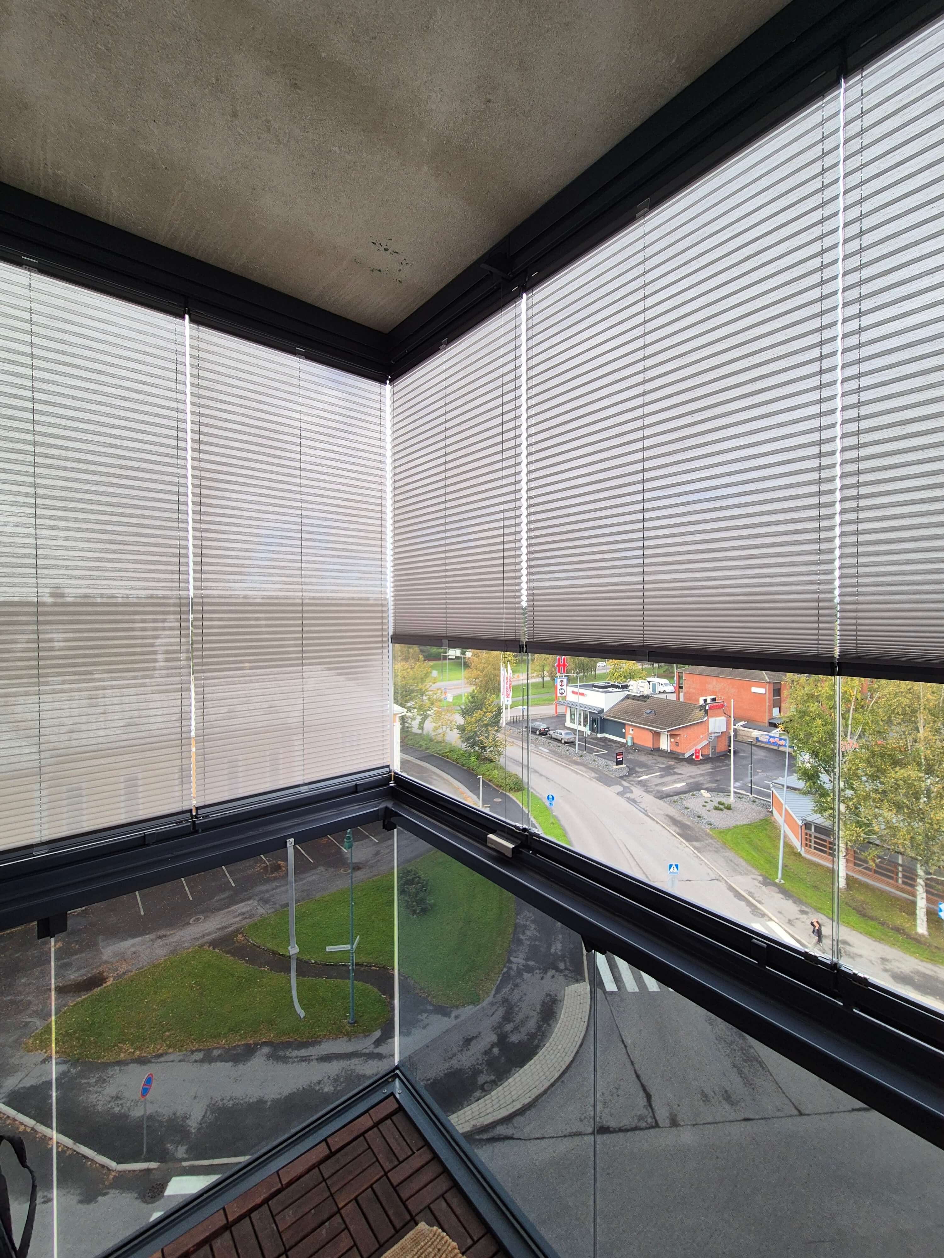 Näkymä parvekkeelta, jossa ritilämatto ja valkoiset sälekaihtimet osittain suljettuina, ulkona katu, rakennuksia ja vihreää nurmikkoa.