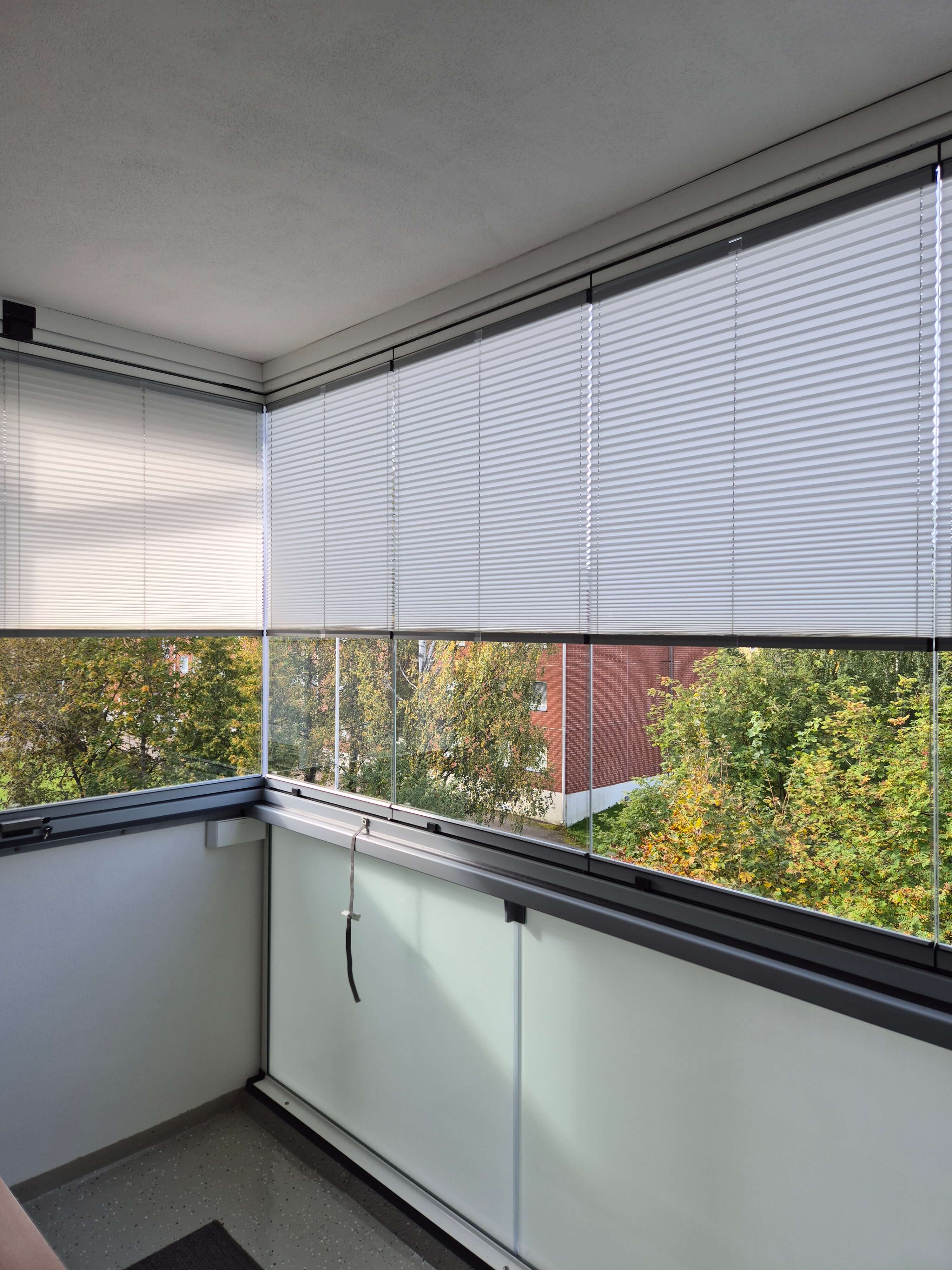 Parvekkeen kulma, jossa on valkoiset sälekaihtimet ja näkymä vihreisiin puihin ja tiilitaloon.