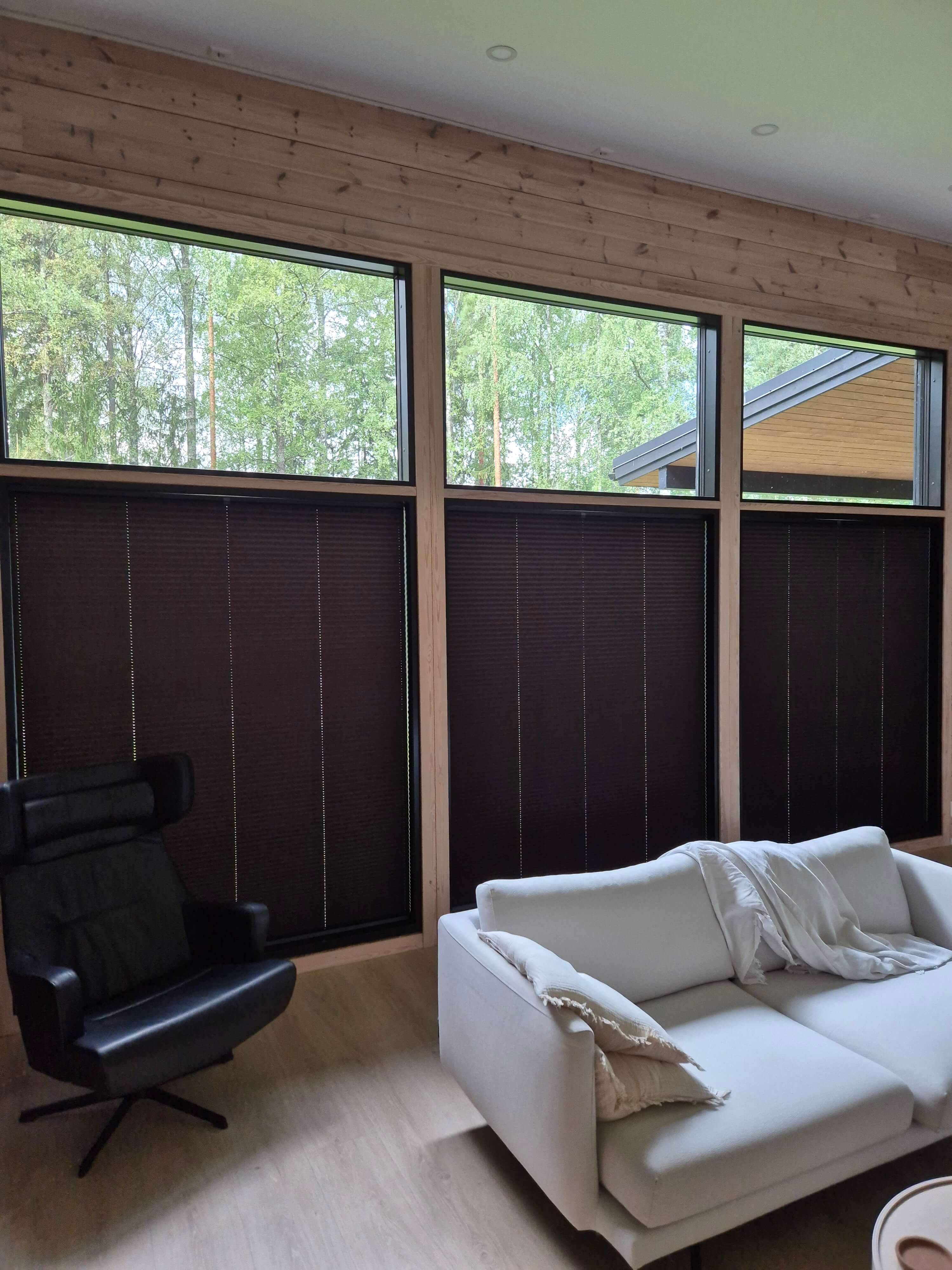 Moderni olohuone, jossa valkoinen sohva tyynyillä ja torkkupeitolla sekä musta nahkainen nojatuoli suuren puisten kehysten ikkunoiden edessä, joiden yläosassa paljaat lasit ja alaosassa pimennetyt kaihtimet.